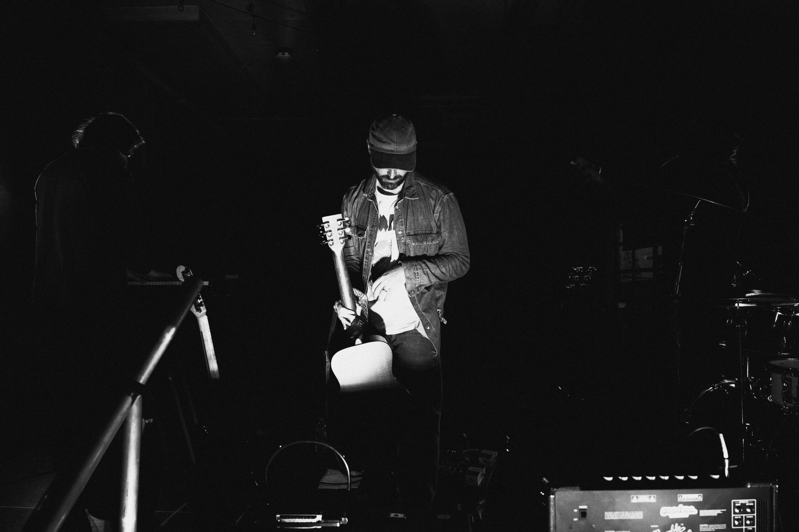 Person wearing a beanie and jacket, holding a drink, standing in a dark setting with musical instruments nearby.