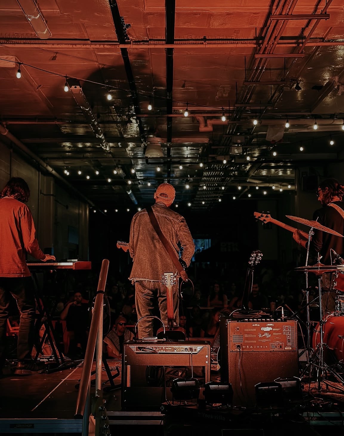 Musicians perform on stage with instruments under warm lighting, audience visible in the background.