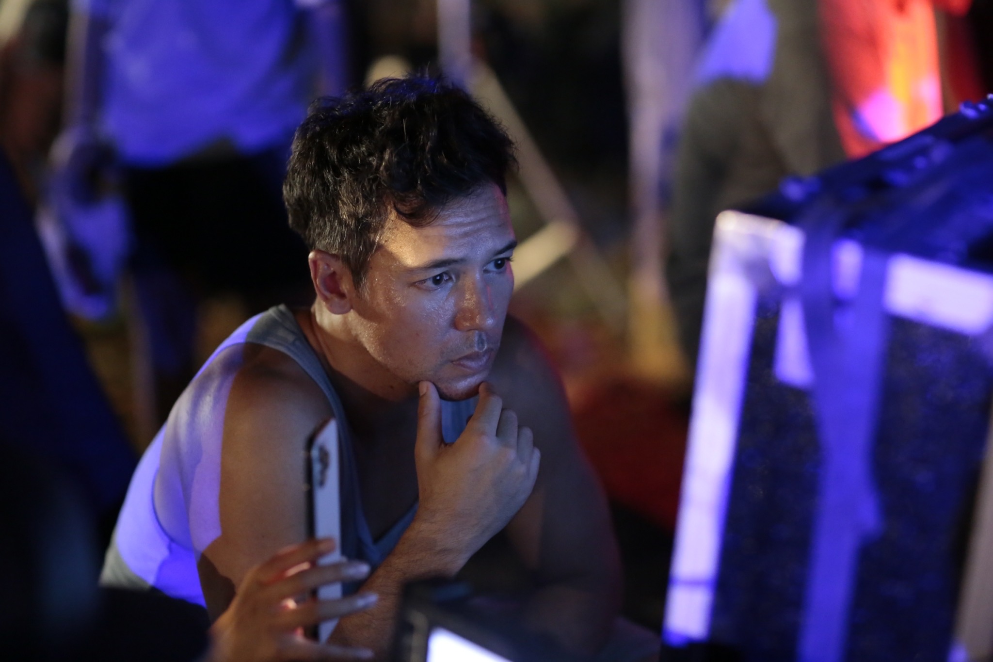 Young man with short dark hair looking at a screen, resting his chin on his hand, in a dimly lit environment