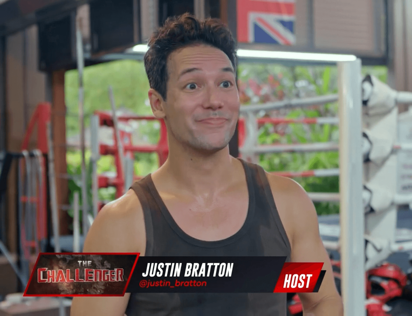 Man with curly hair in a tank top smiling indoors with gym equipment in background.