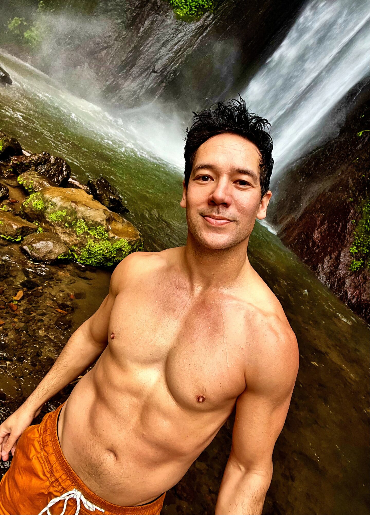 Man with short dark hair and a muscular torso stands near a waterfall, smiling, with water and rocks in background.