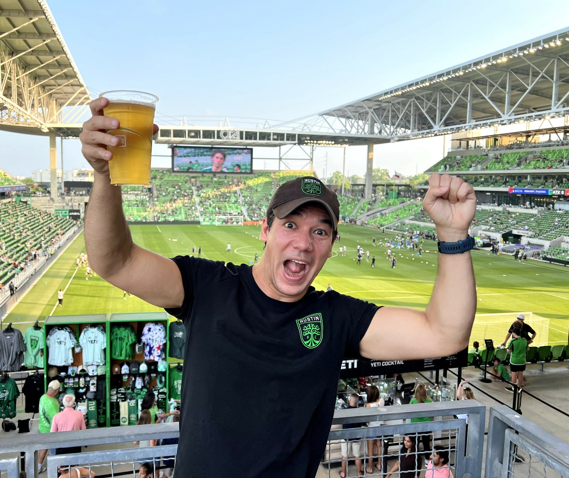 Person celebrating with a raised glass at a sports stadium, cheering with a clenched fist, crowd and field in background.