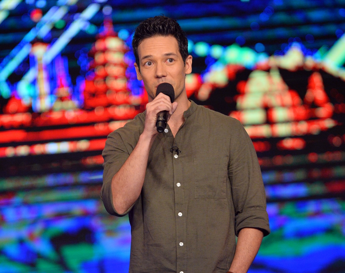 Man holding microphone on stage with colorful, blurred background lights, wearing a green shirt, looking at camera.