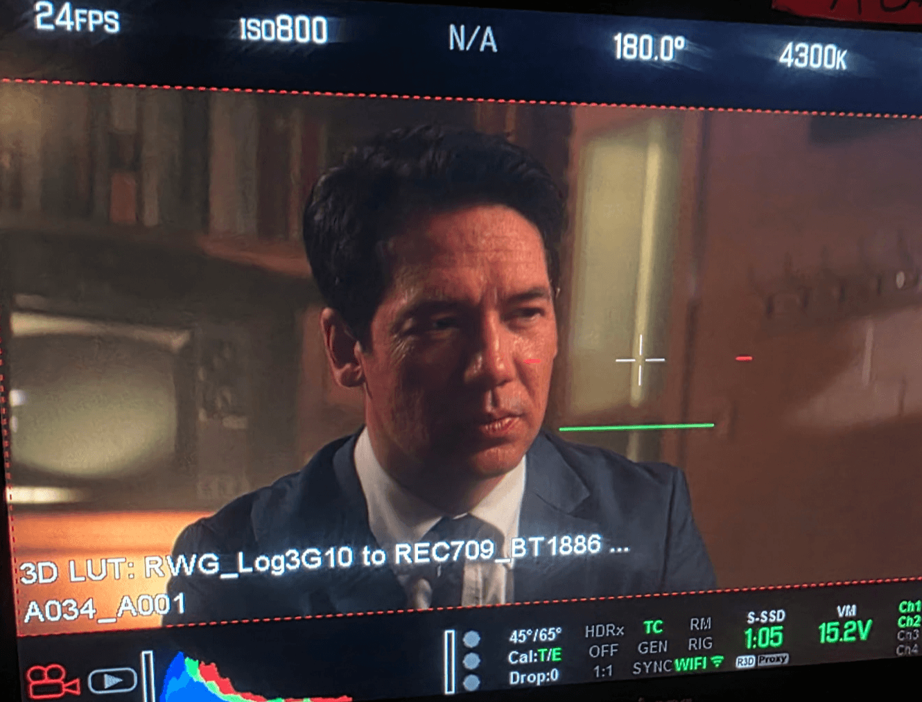 Man with dark hair in a suit looks to the side in a room with bookshelves, on a film or video set