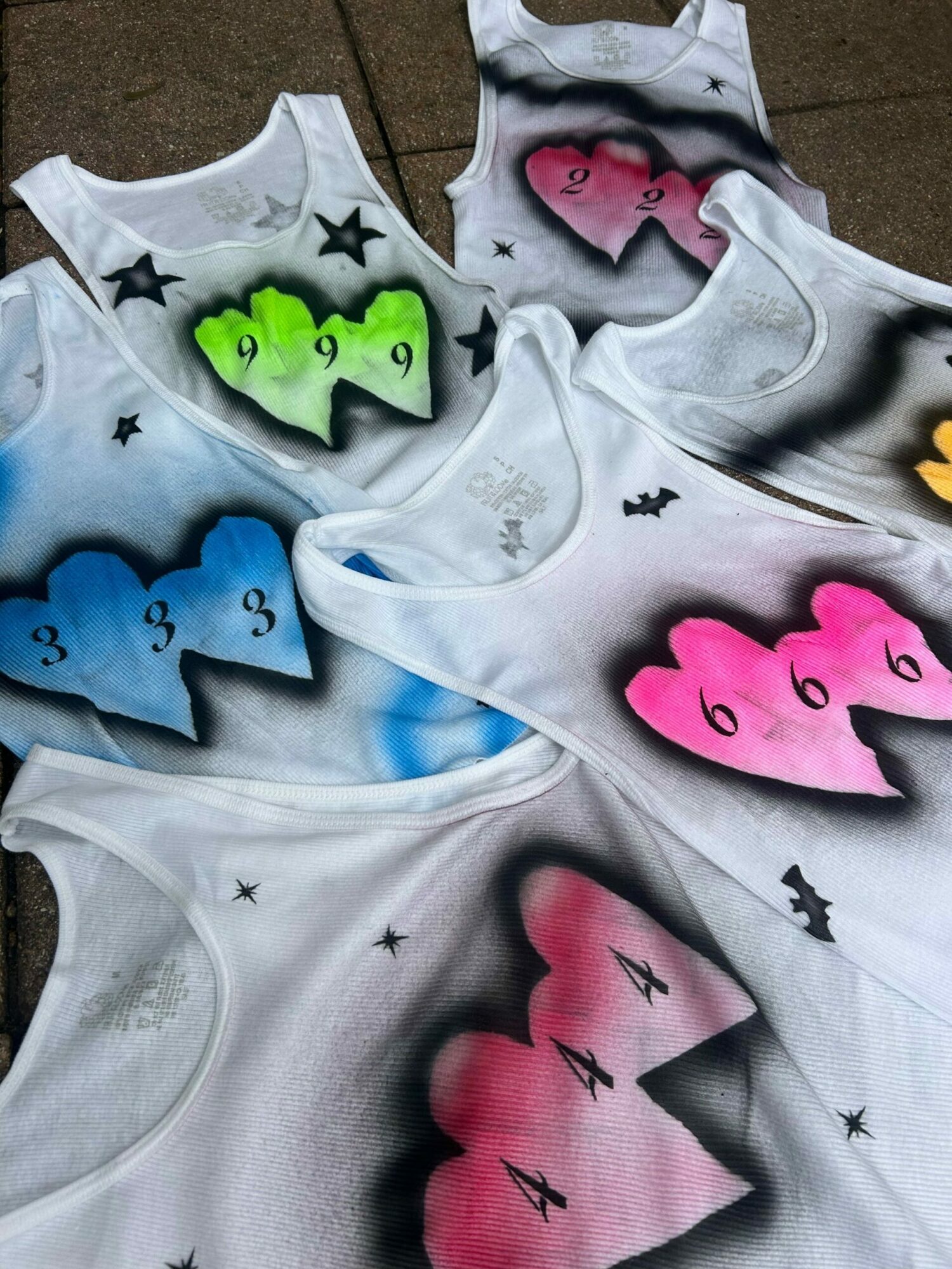 White T-shirts with colorful, cartoonish cloud and star designs, some with numbers and bat illustrations, laid out on a surface.