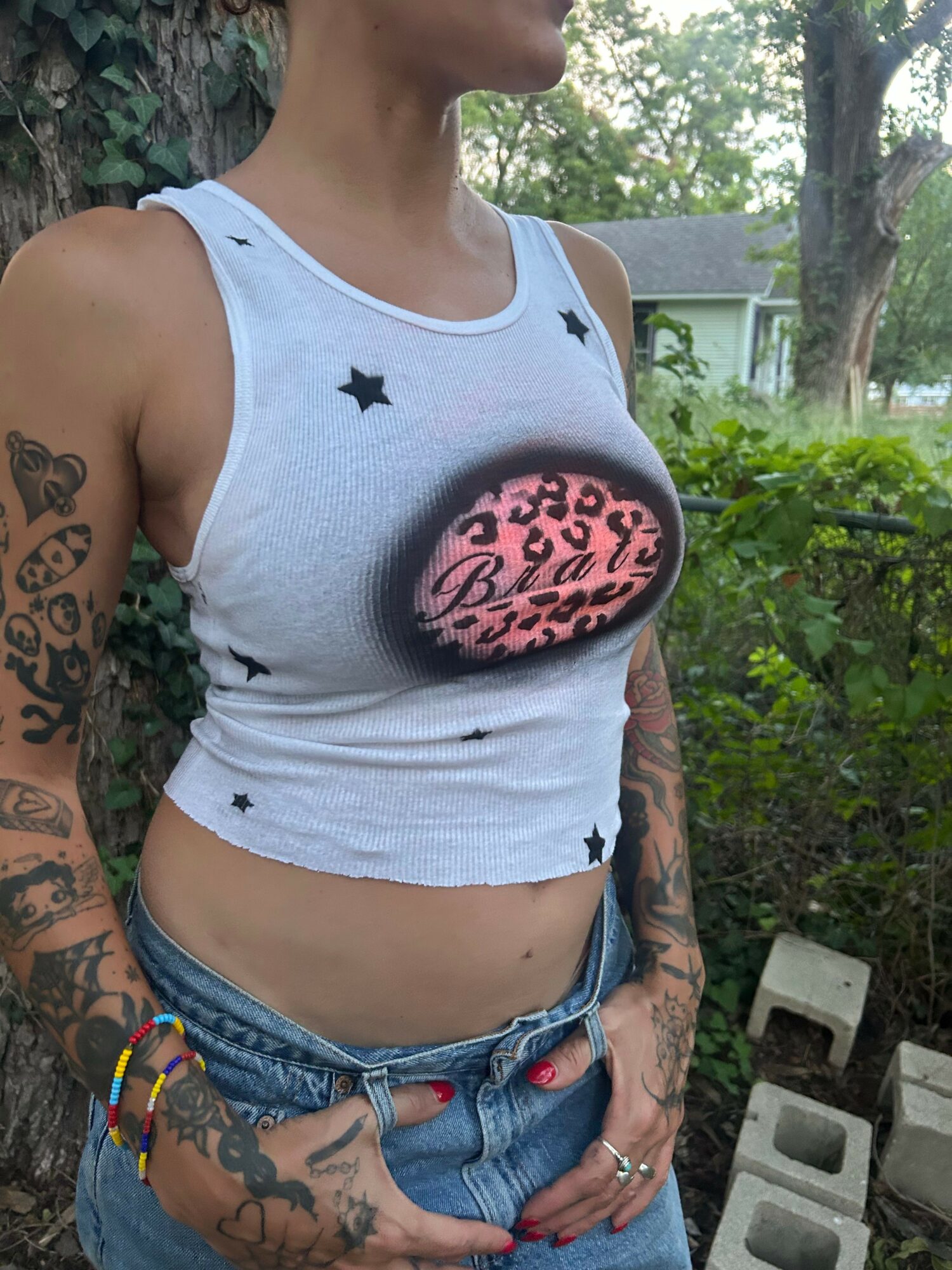 Person with tattoos wearing a white tank top and denim shorts outdoors, with trees and greenery in background.