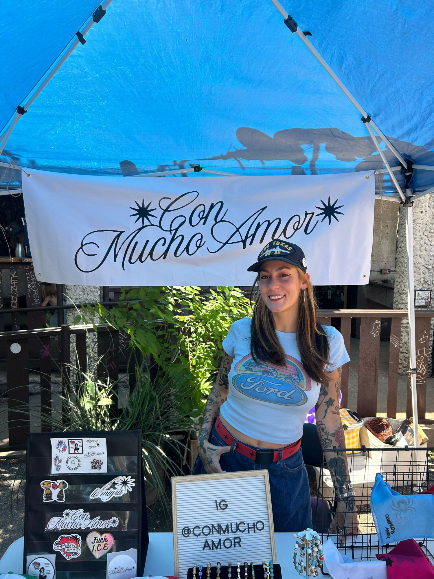 Woman standing outdoors near a blue tent with a sign that reads 'Maui Hemp' and a smaller sign with information, surrounded by plants.