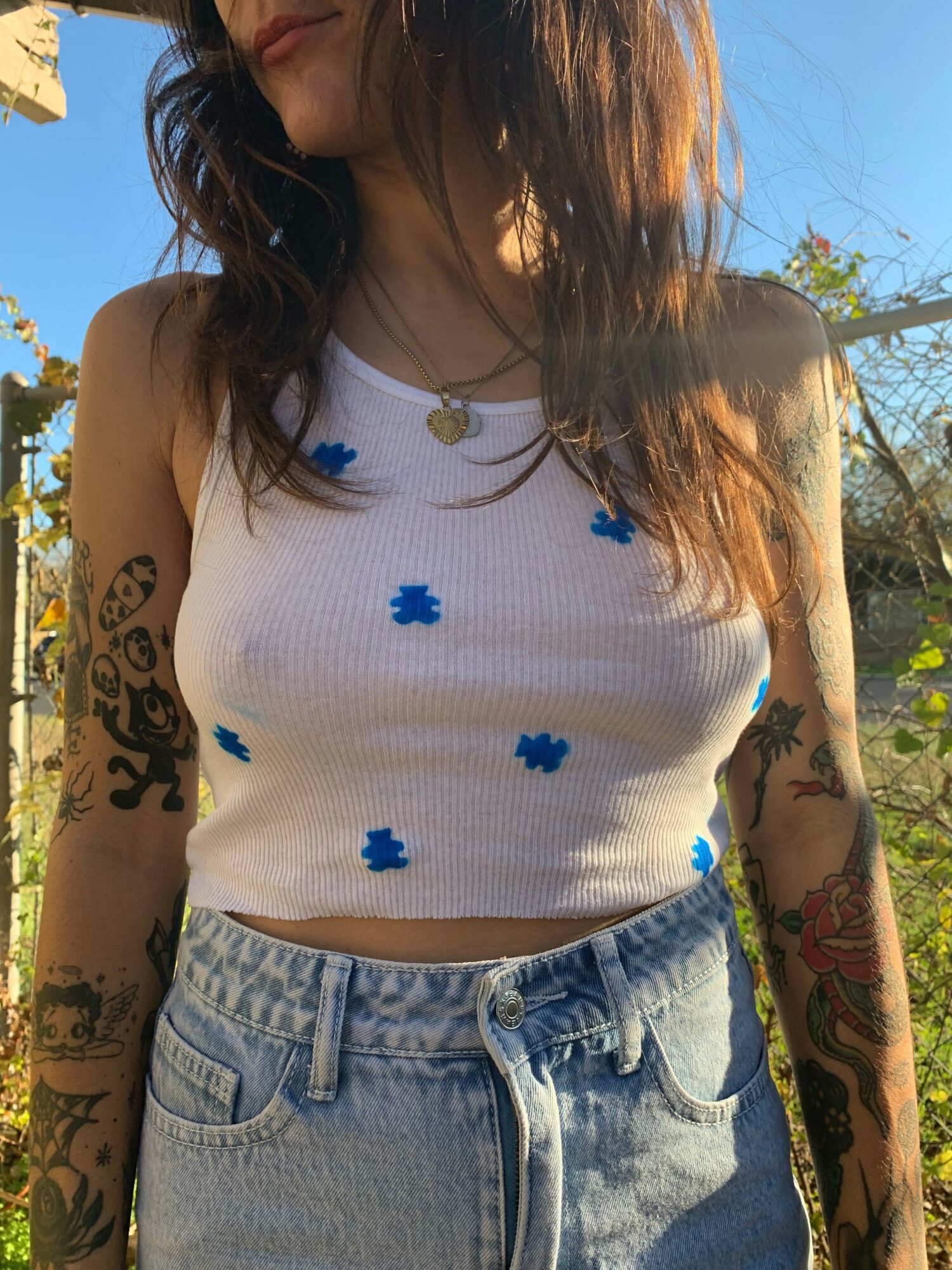 Person with tattoos on arms wearing a white top with blue flower patterns and denim jeans outdoors.