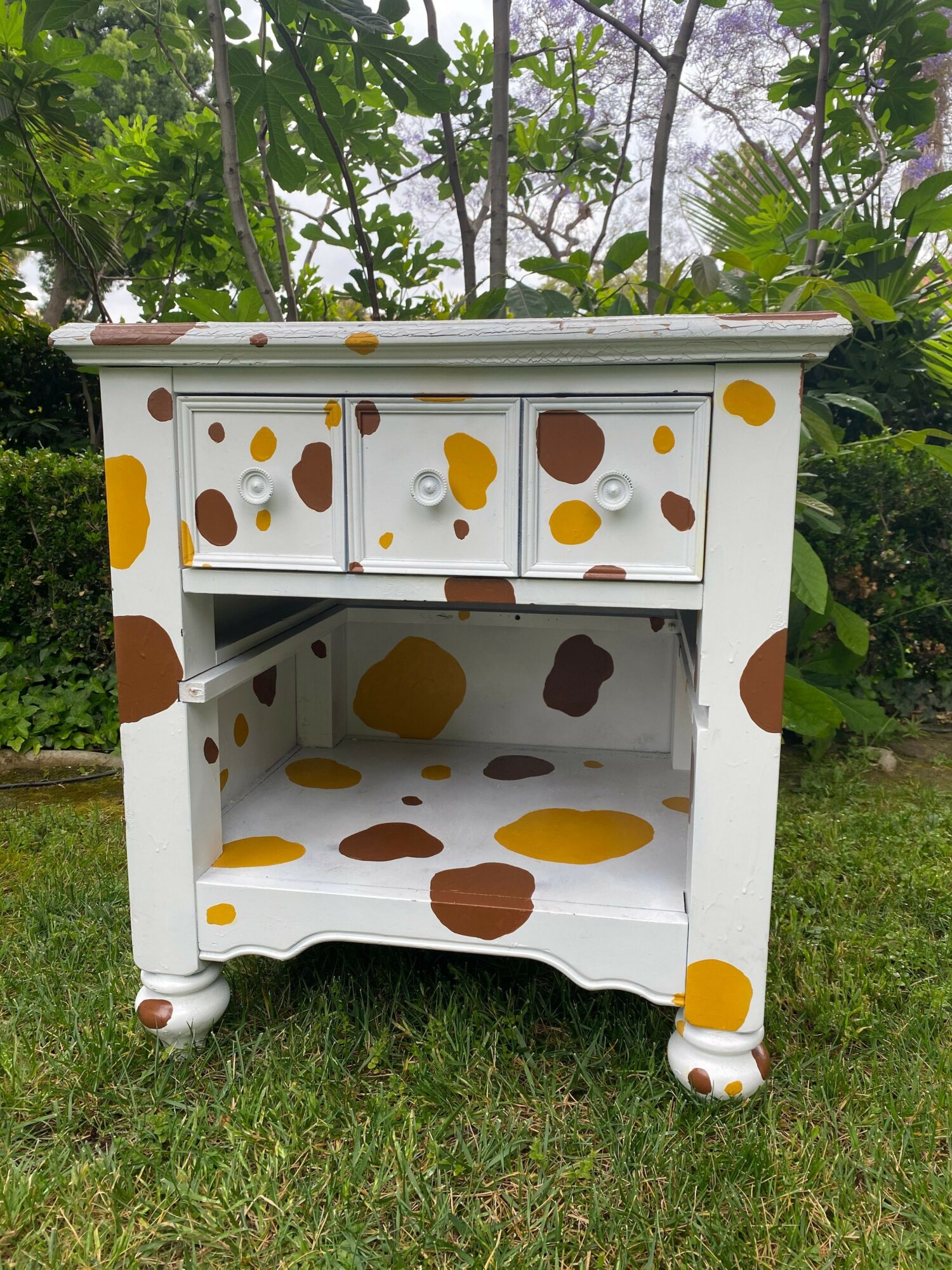 White children's playhouse with brown and yellow spots, open door, surrounded by grass and trees.