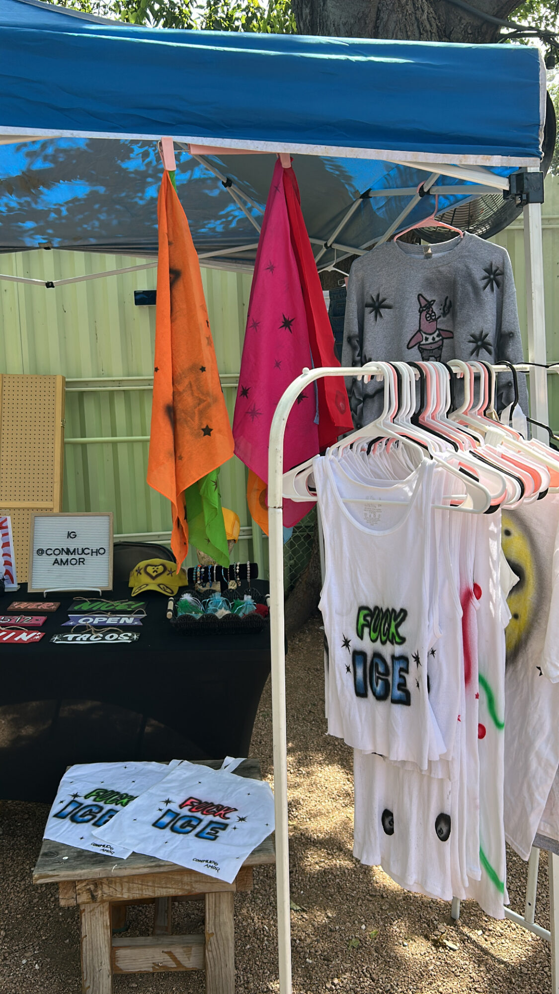 Outdoor market stall with colorful clothing and accessories, green fence in background, sunlight, and various items displayed.
