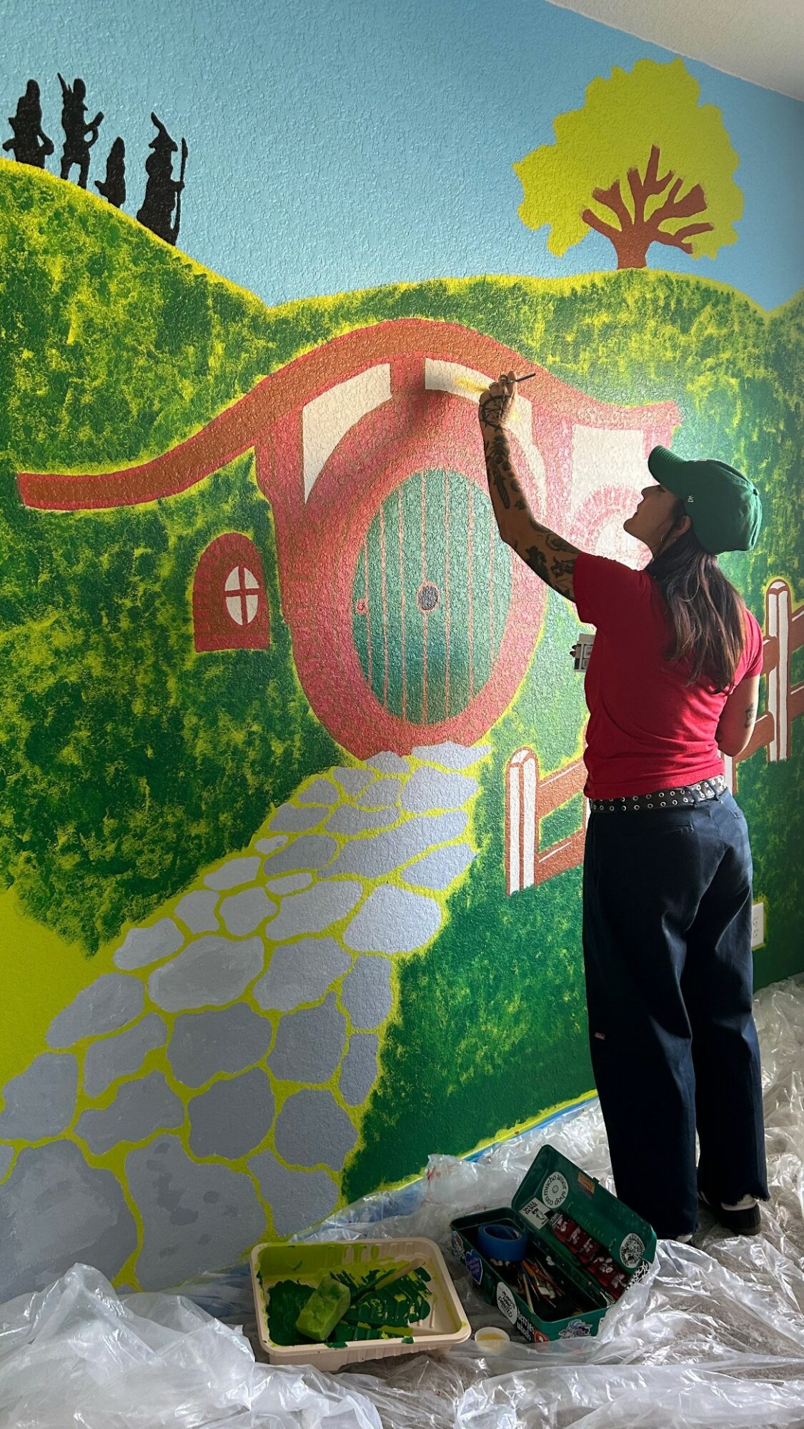 Person painting a colorful mural of a house, pathway, and trees on a wall, with painting supplies on the floor.