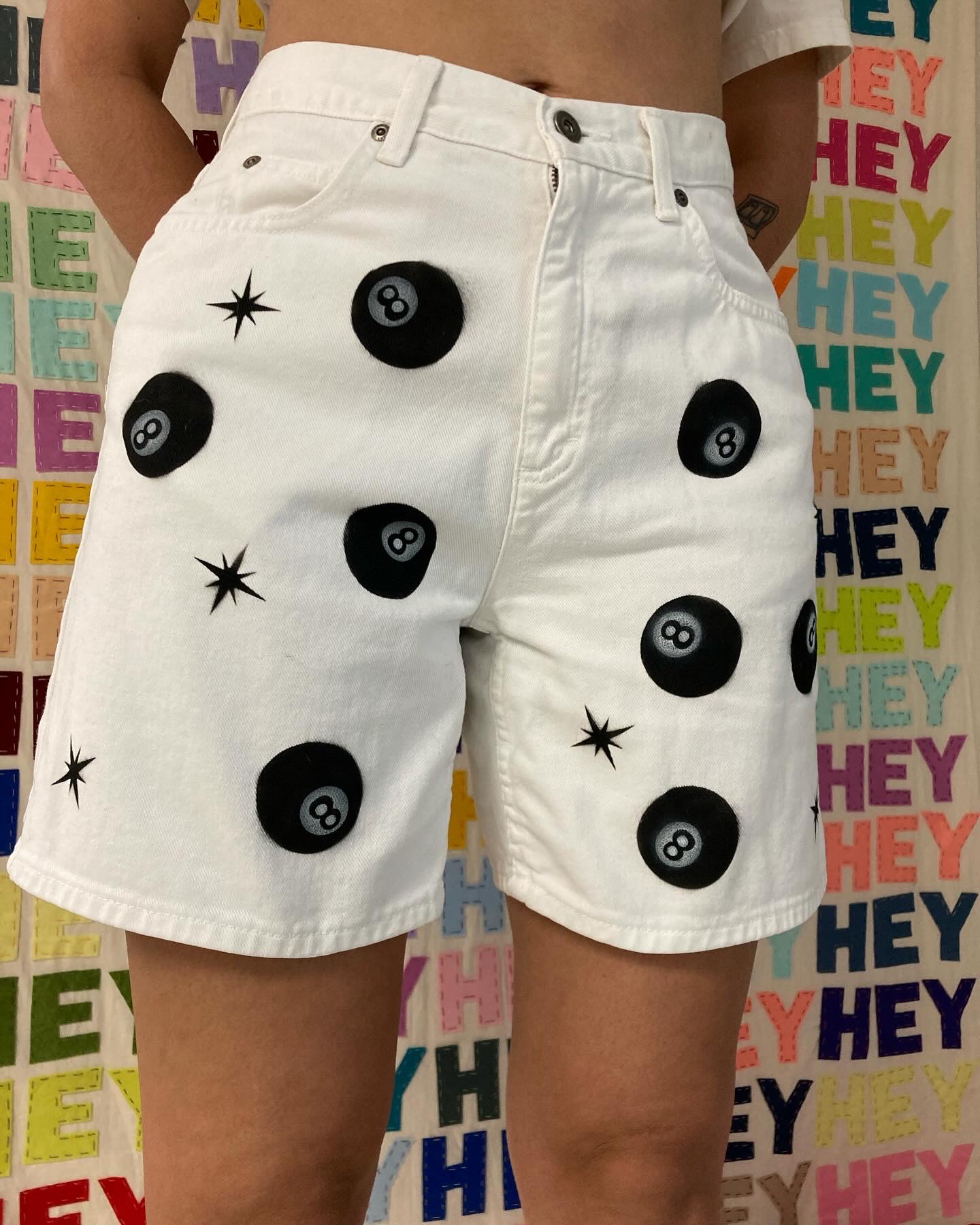 Person wearing white shorts with black buttons and star-shaped embellishments, standing in front of a colorful background with repeated text.