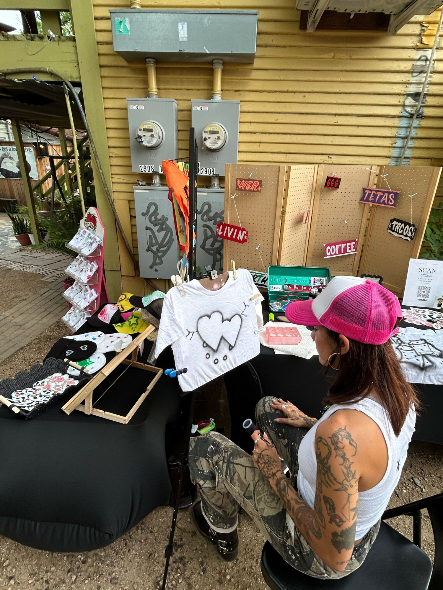 Person with tattoos wearing a pink cap drawing on a white canvas at an outdoor event, with art supplies and decorations on table.