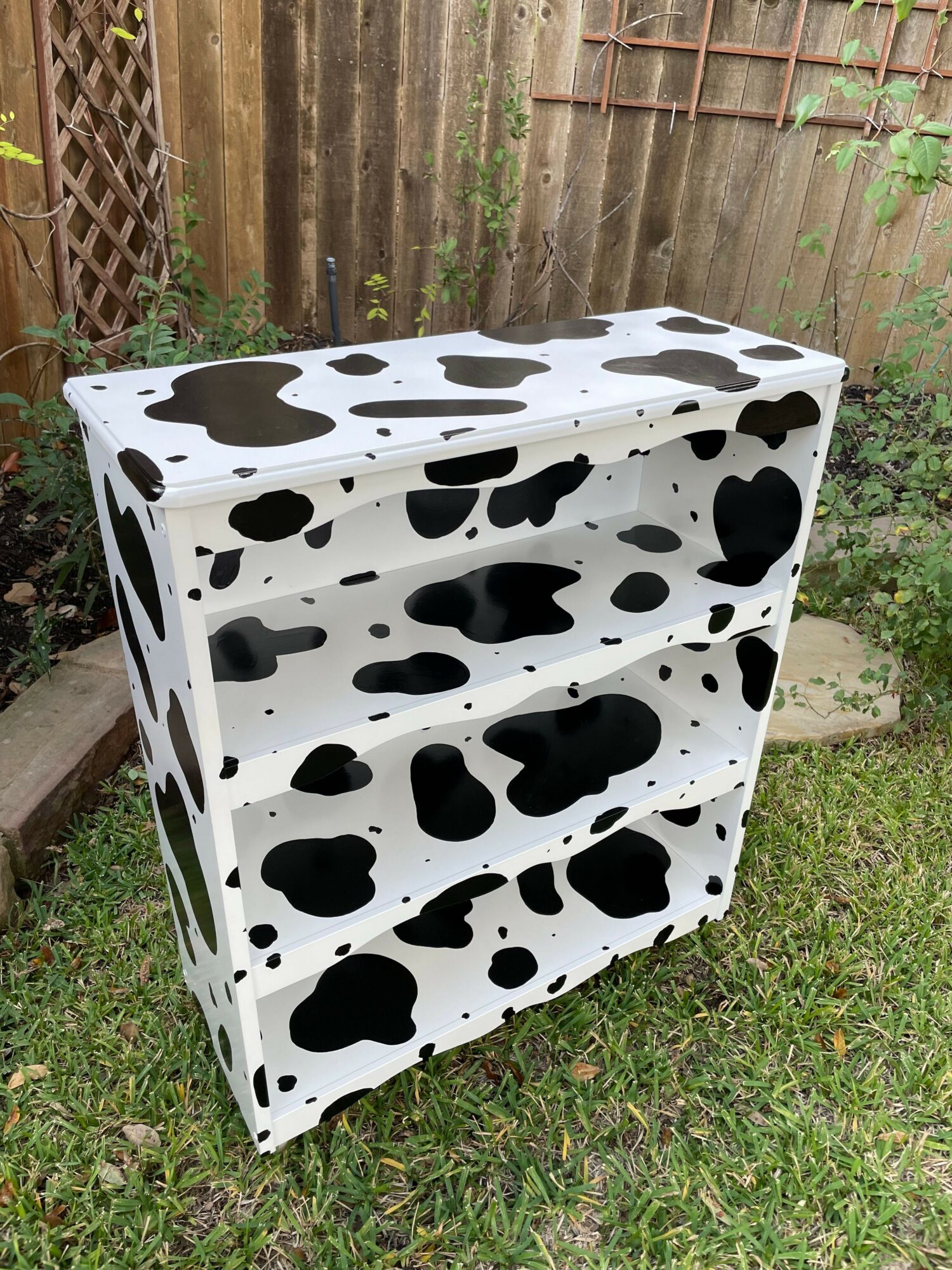 A white rectangular object with black cow-like spots, placed outdoors on grass with a wooden fence in the background.