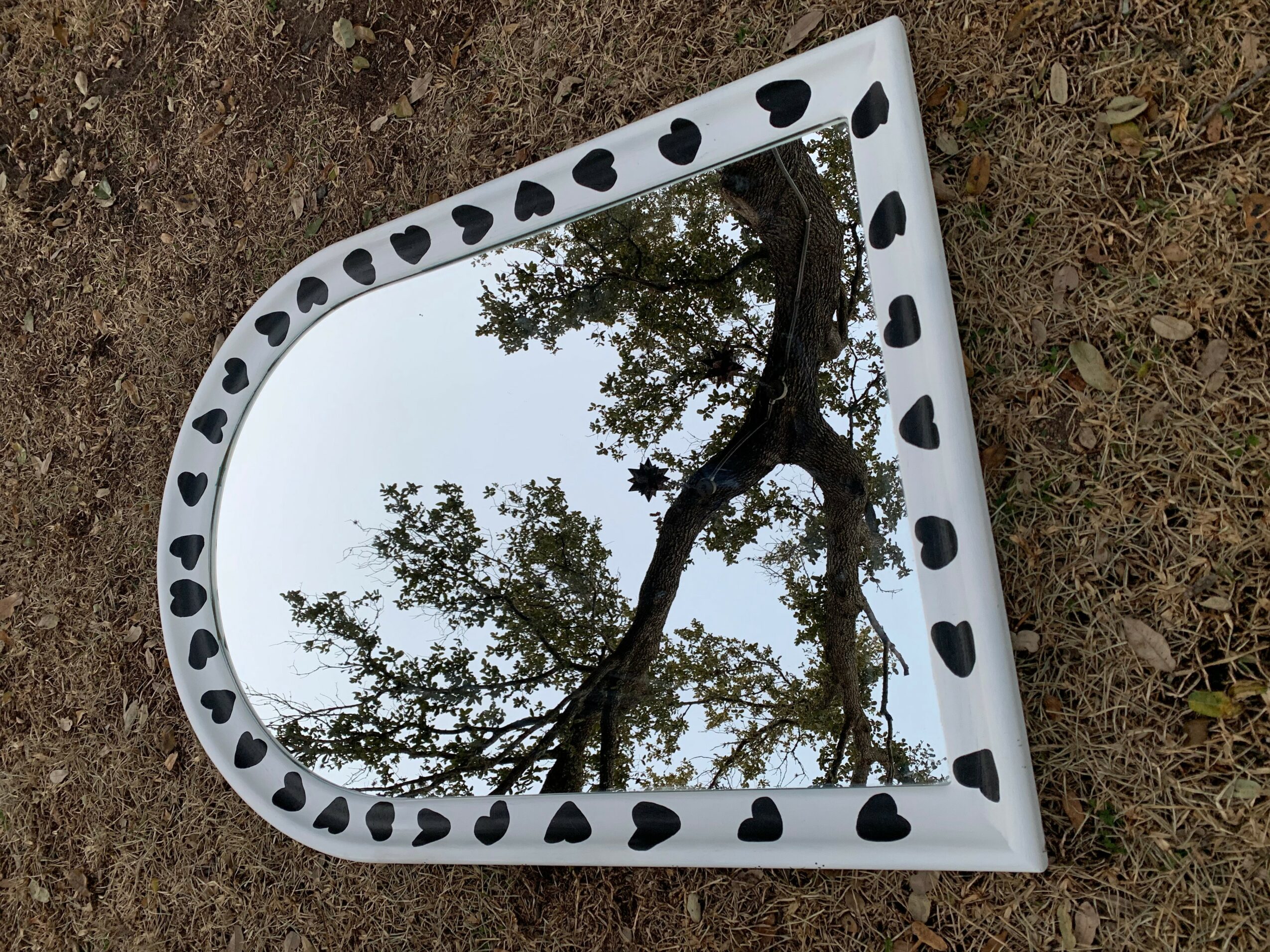 A mirror with a white frame and black heart-shaped decorations, reflecting a tree and sky.