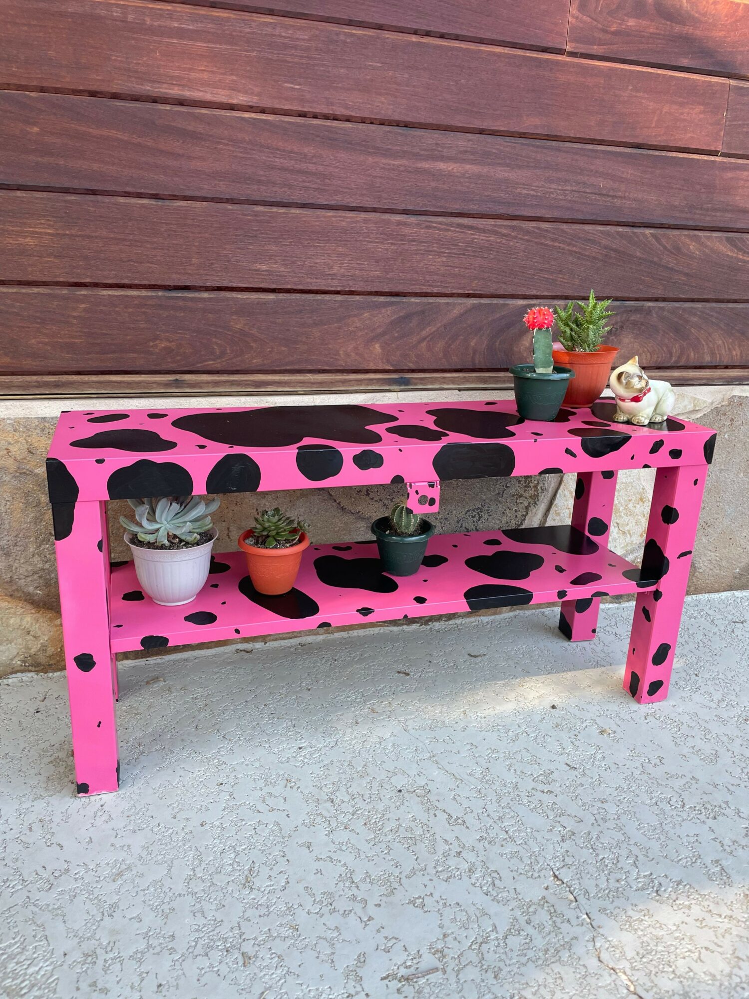 Pink wooden bench with black spots, holding four small potted plants, on a wooden and concrete surface.