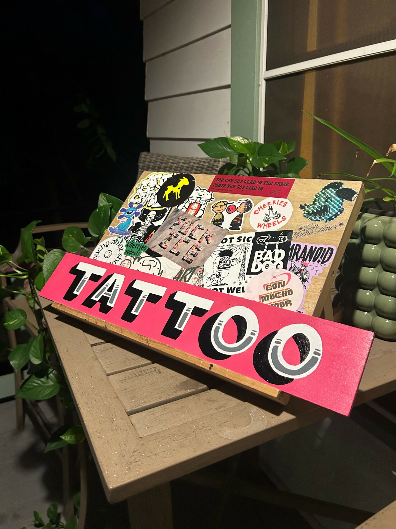 Pink sign with black and white text reading 'TATTOO' on a table with various stickers and a plant.