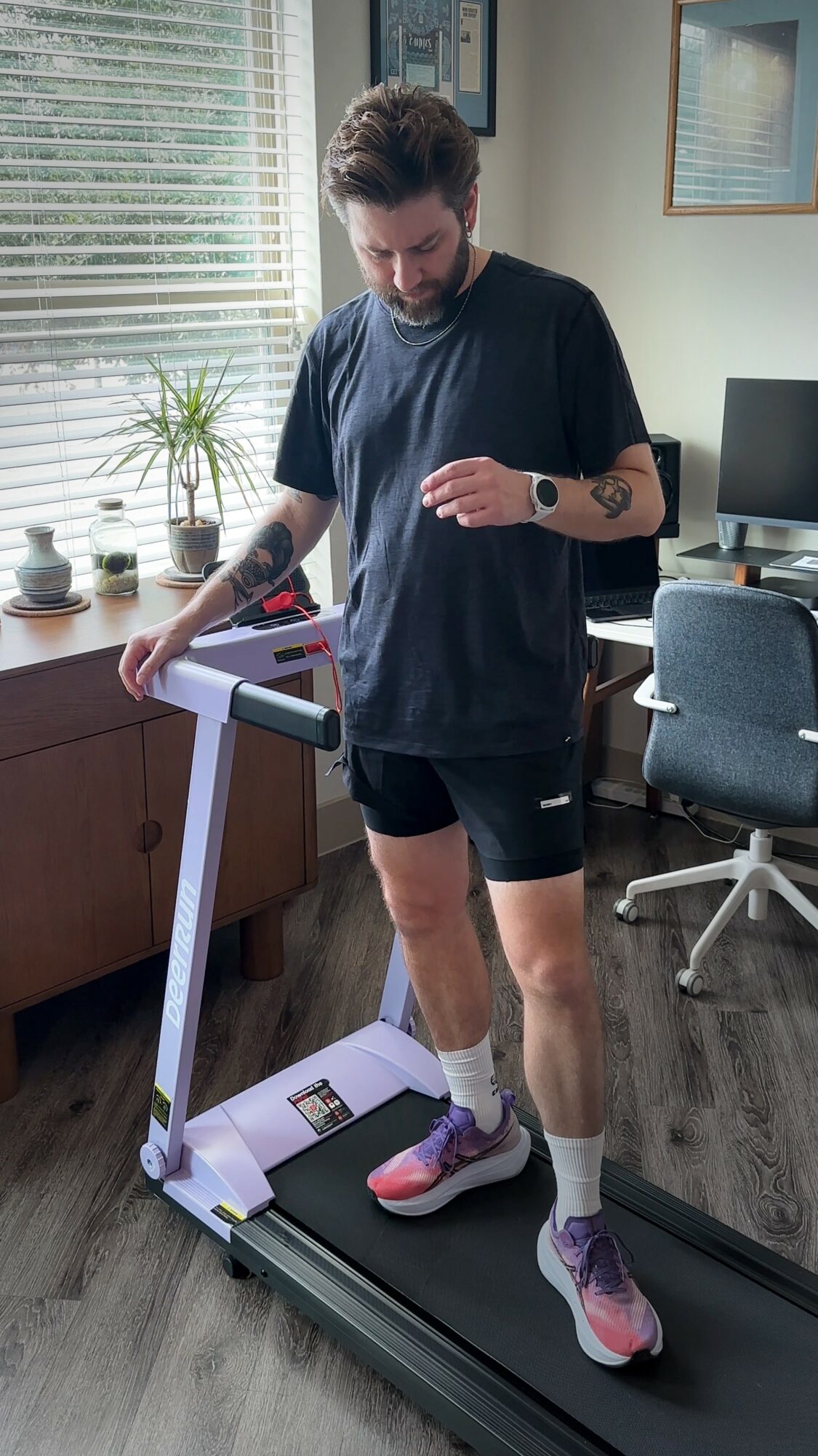 Person standing on a treadmill in a room with a desk, chair, and window, wearing athletic clothes and looking down.