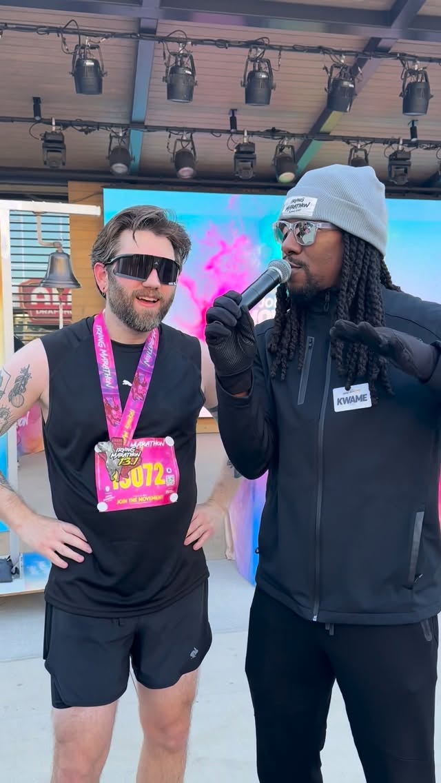 Two men on stage, one with sunglasses and a medal, the other with a beanie and microphone, in front of a screen.