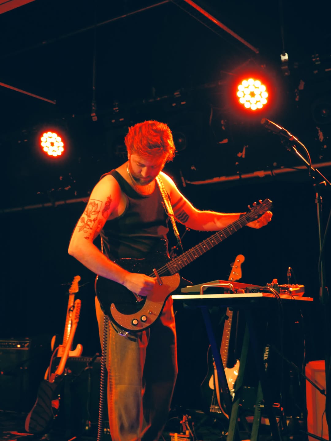 Musician playing an electric guitar on stage with red stage lights, wearing a sleeveless shirt, tattoos visible on arms.