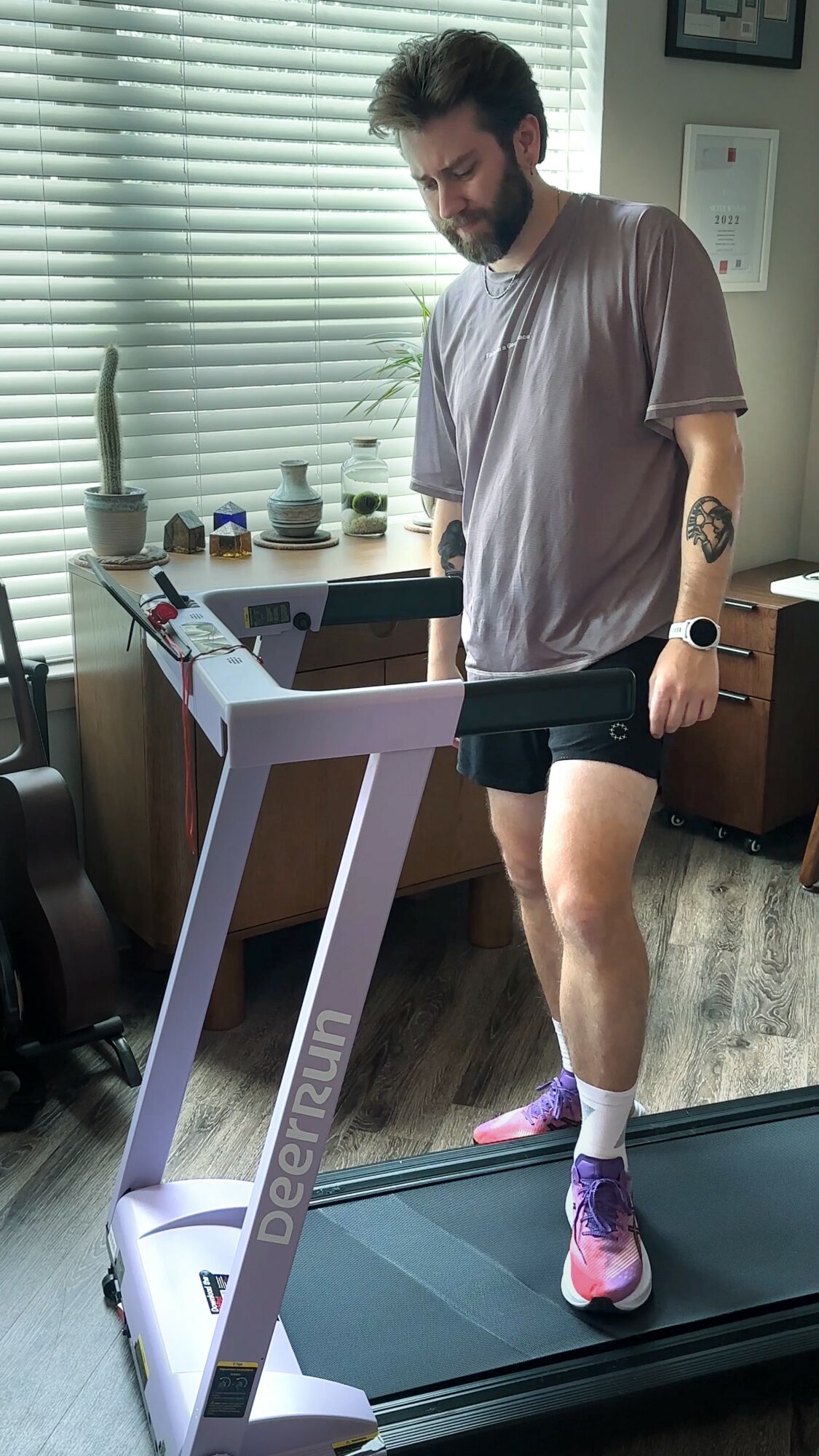 Man walking on a treadmill in a room with window blinds, wearing a gray t-shirt, black shorts, and purple shoes.