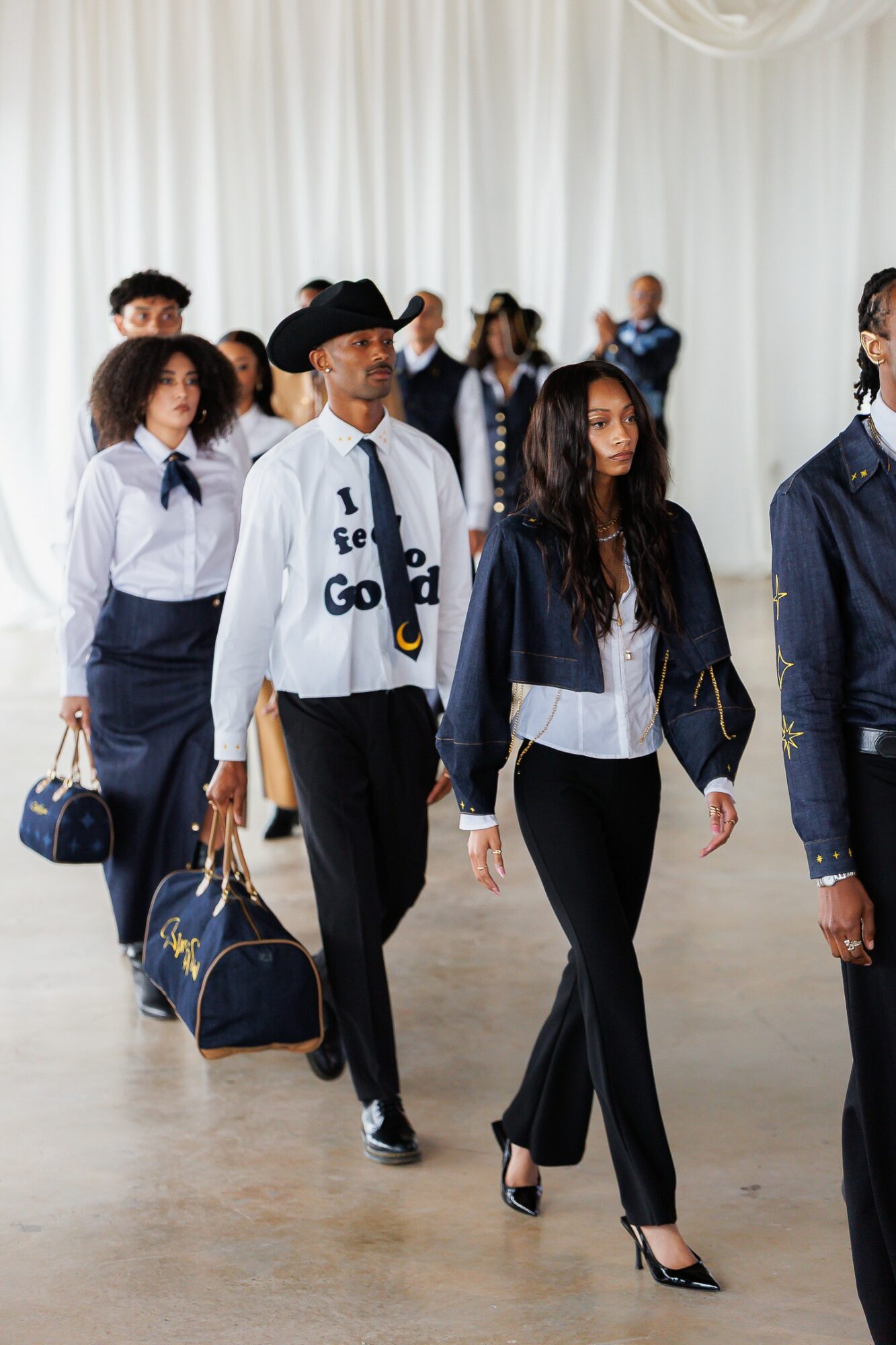 Group of people walking indoors, some carrying bags, with a white curtain background.