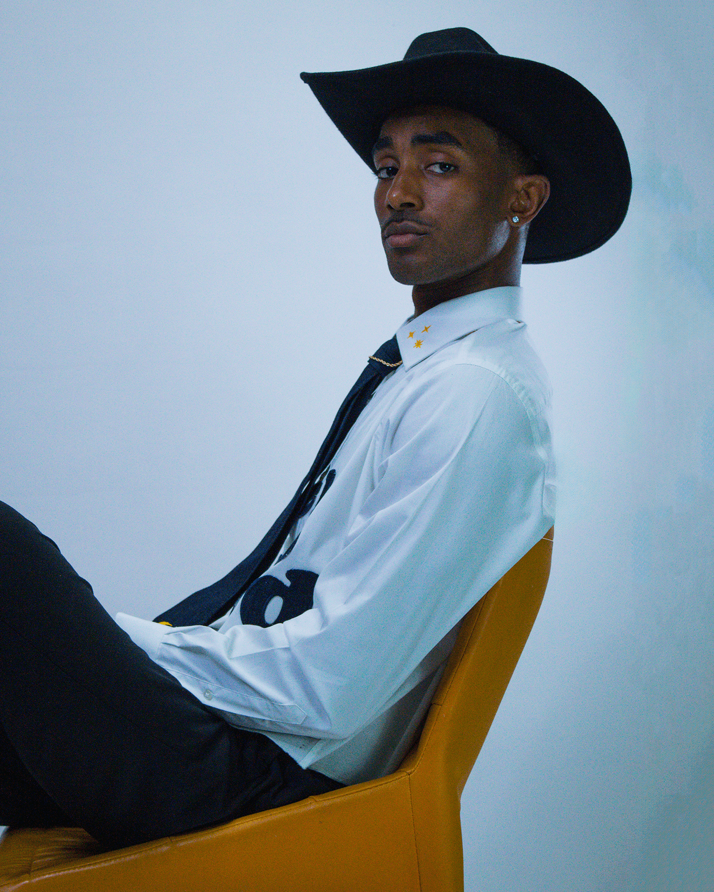 Young man wearing a large black hat, white shirt, and black tie, sitting sideways in a yellow chair against a plain background.