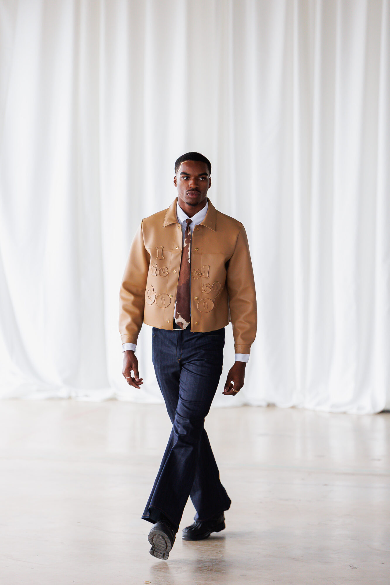 Person walking on runway with white curtain background, wearing a tan jacket, dark pants, and a patterned shirt.