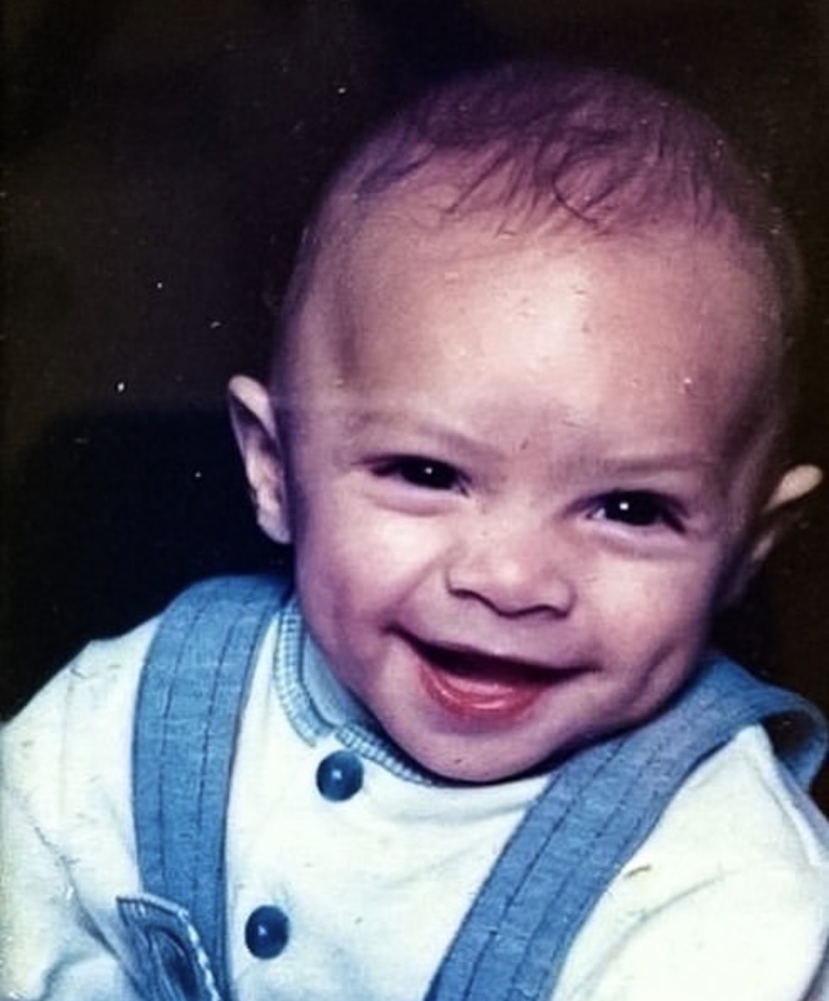 Smiling baby with short hair wearing a white shirt and blue suspenders.