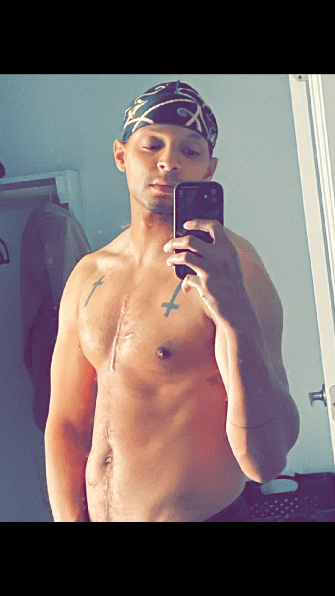 Young man taking a mirror selfie, shirtless, wearing a bandana, with a cross tattoo on chest.