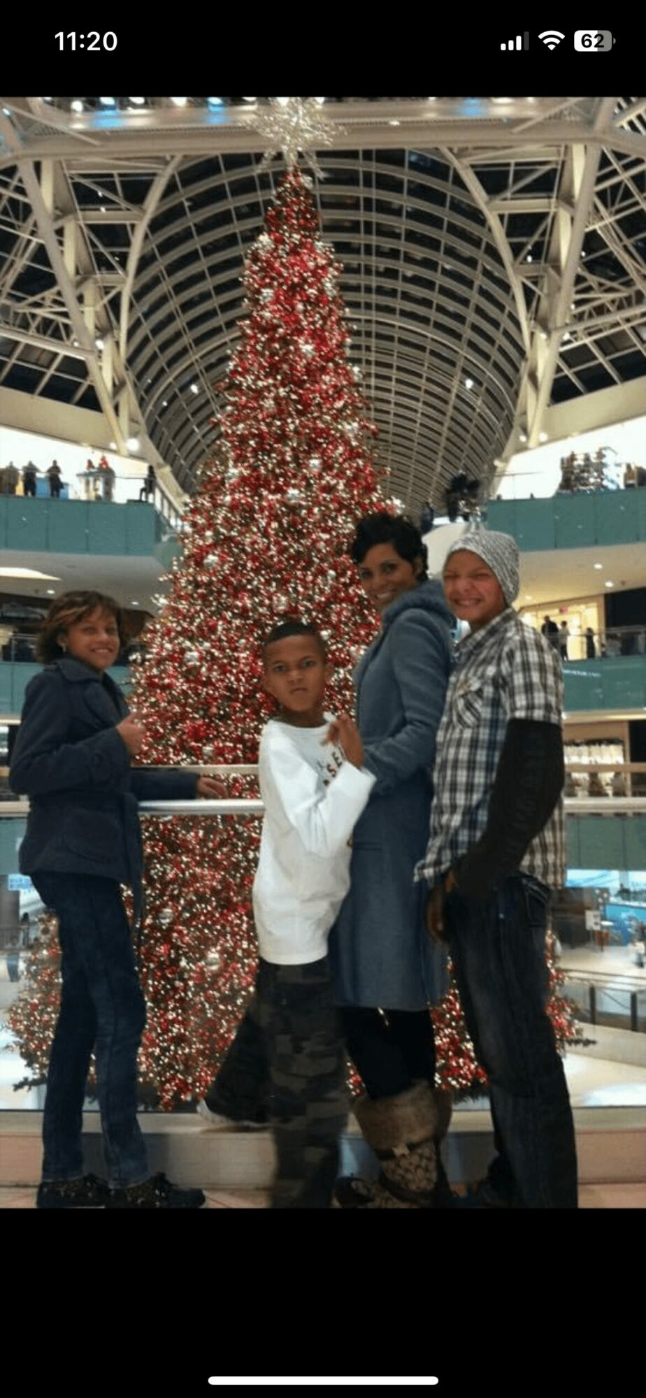 Four people stand in front of a decorated Christmas tree inside a shopping mall, with a glass ceiling above.