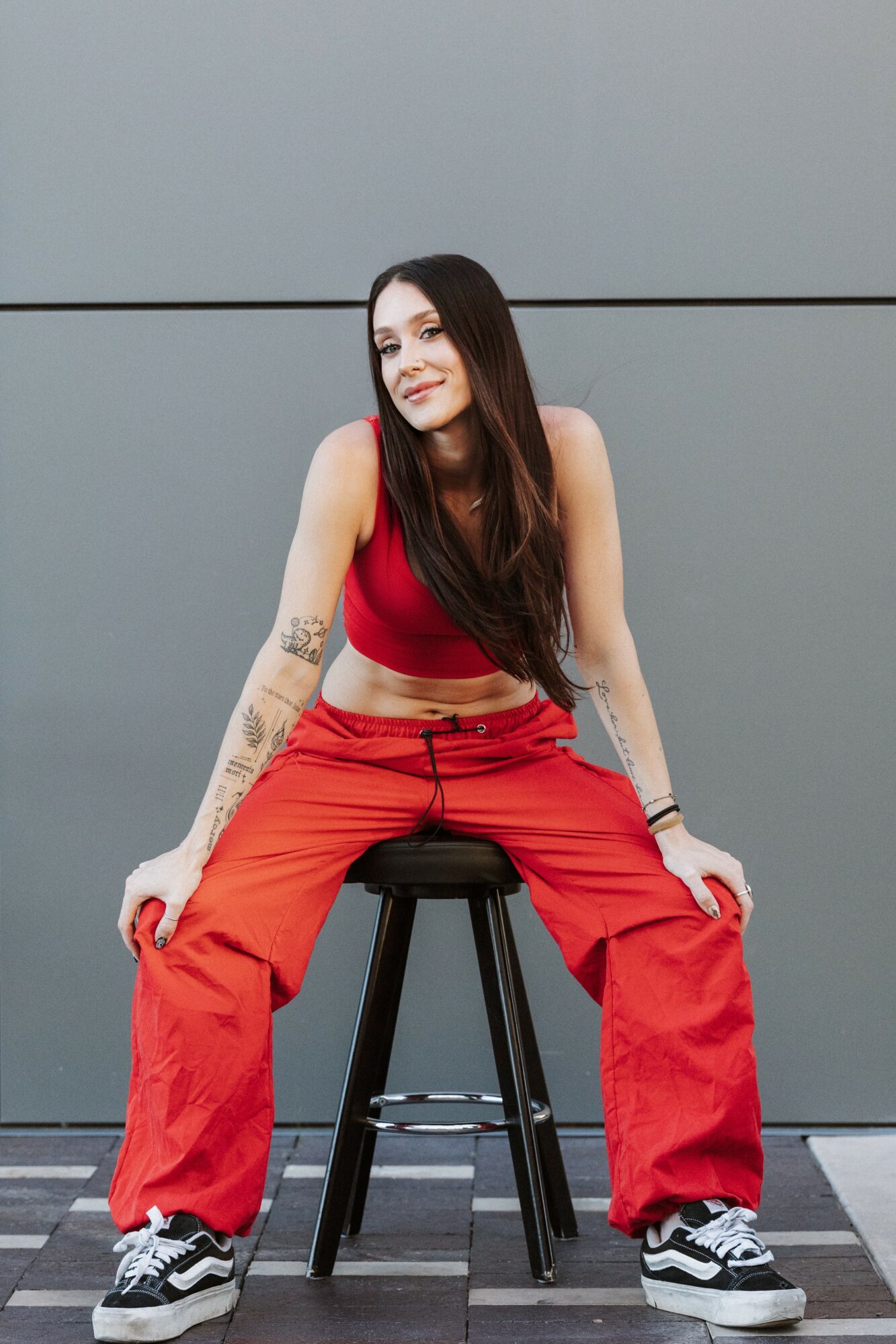 Young woman with long dark hair sitting on a stool against a gray wall, wearing a red crop top and orange pants.