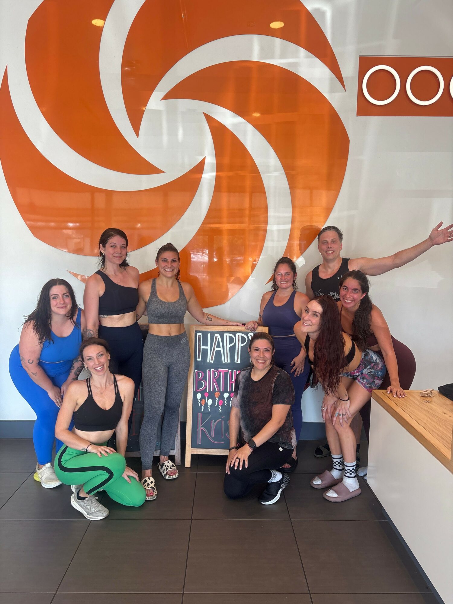 Group of six people posing in front of a large orange and white swirl logo, some sitting and some standing, in a room with dark floor and wall.