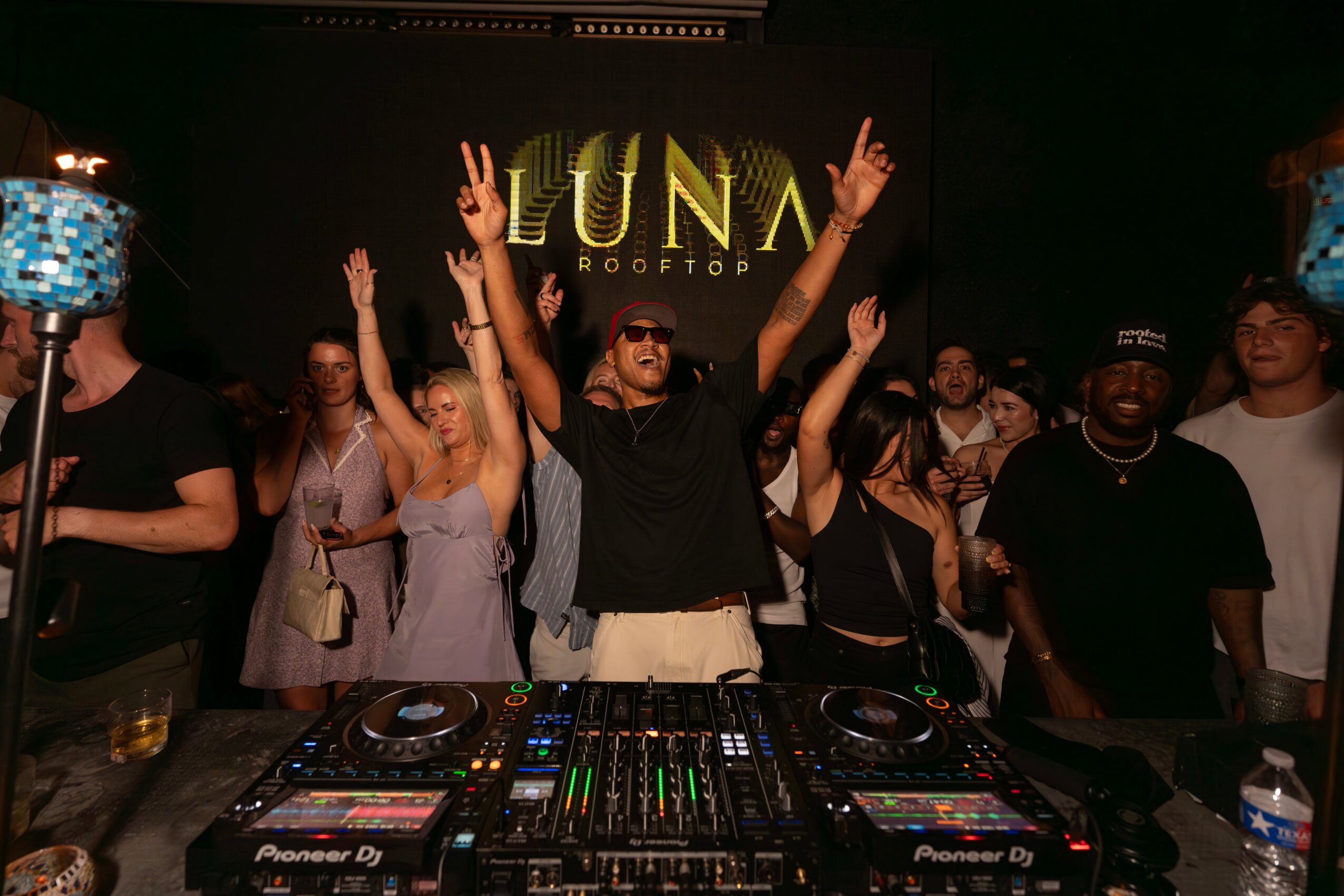 Group of people dancing and raising hands at a party with a DJ setup and a screen displaying 'LUNA ROOFTOP'.