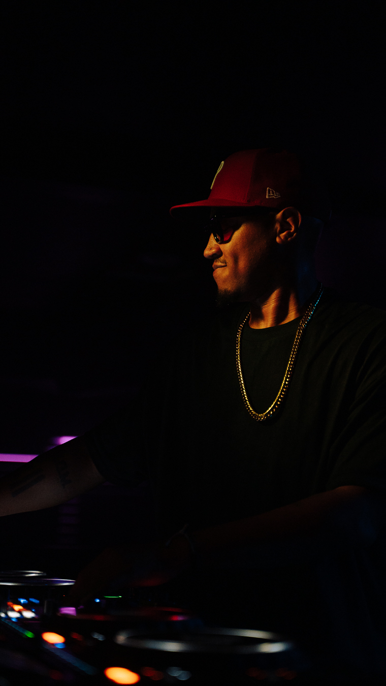 Person wearing a red cap, sunglasses, and a gold chain, illuminated in a dark environment.
