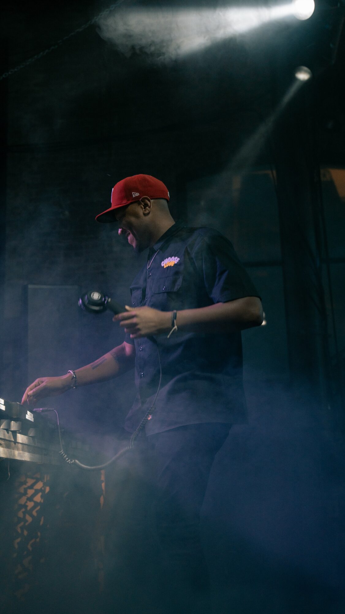 Person wearing a red cap and dark shirt, holding a device, in a dimly lit environment with stage lighting.