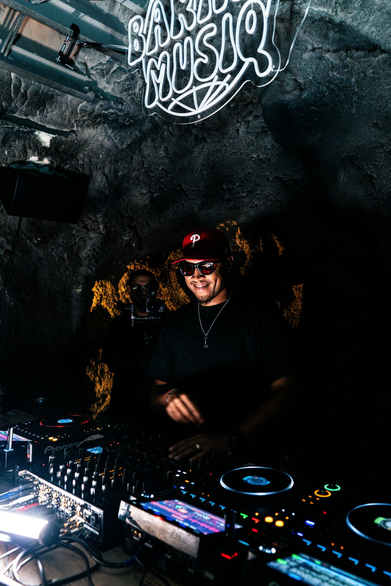 DJ wearing sunglasses and a cap, smiling behind a DJ setup at a music event, with a neon sign above.
