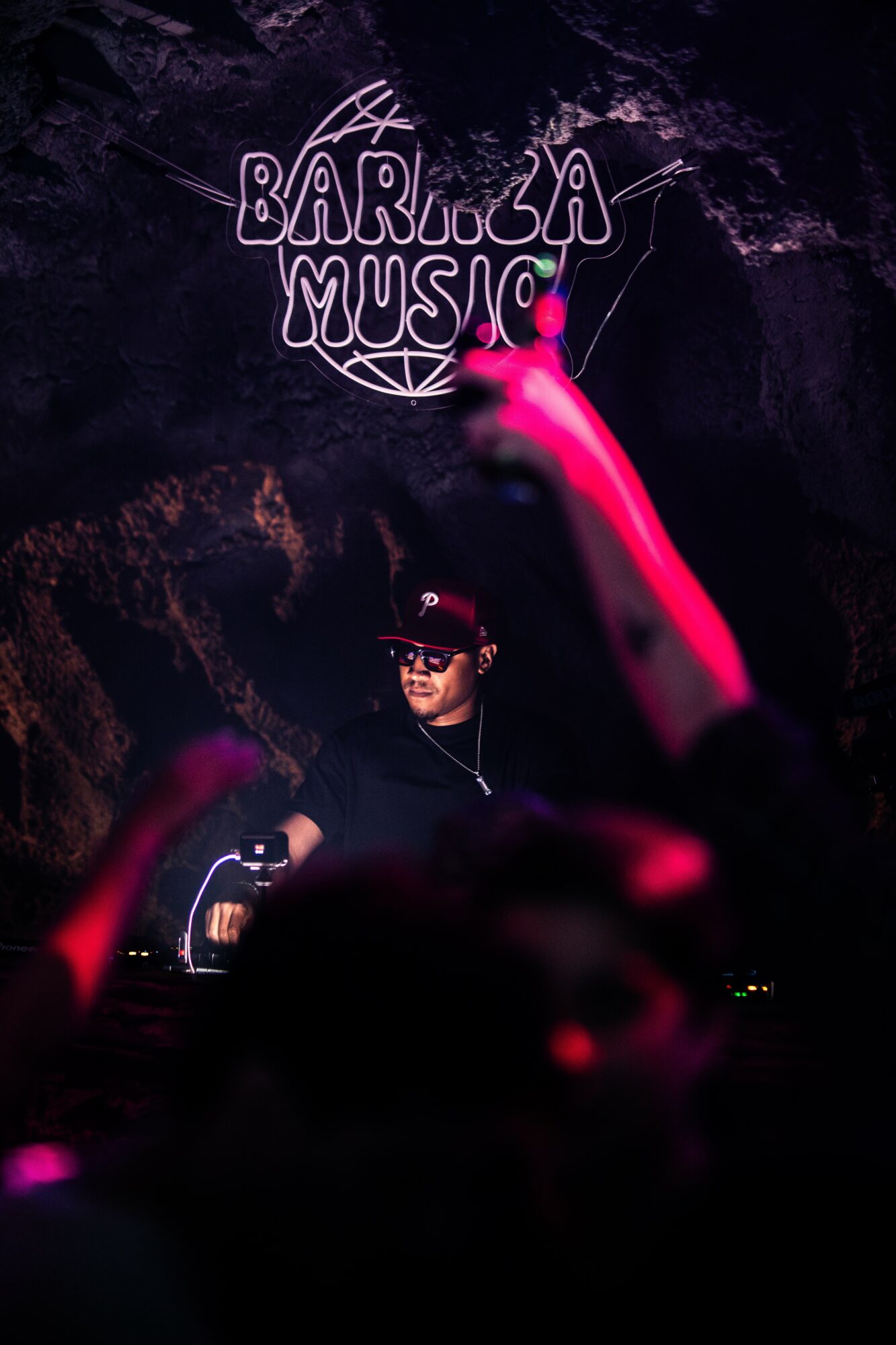 DJ performing at a music event with a neon sign reading 'BARRIO MUSIC' in the background, crowd with raised hands, dark setting