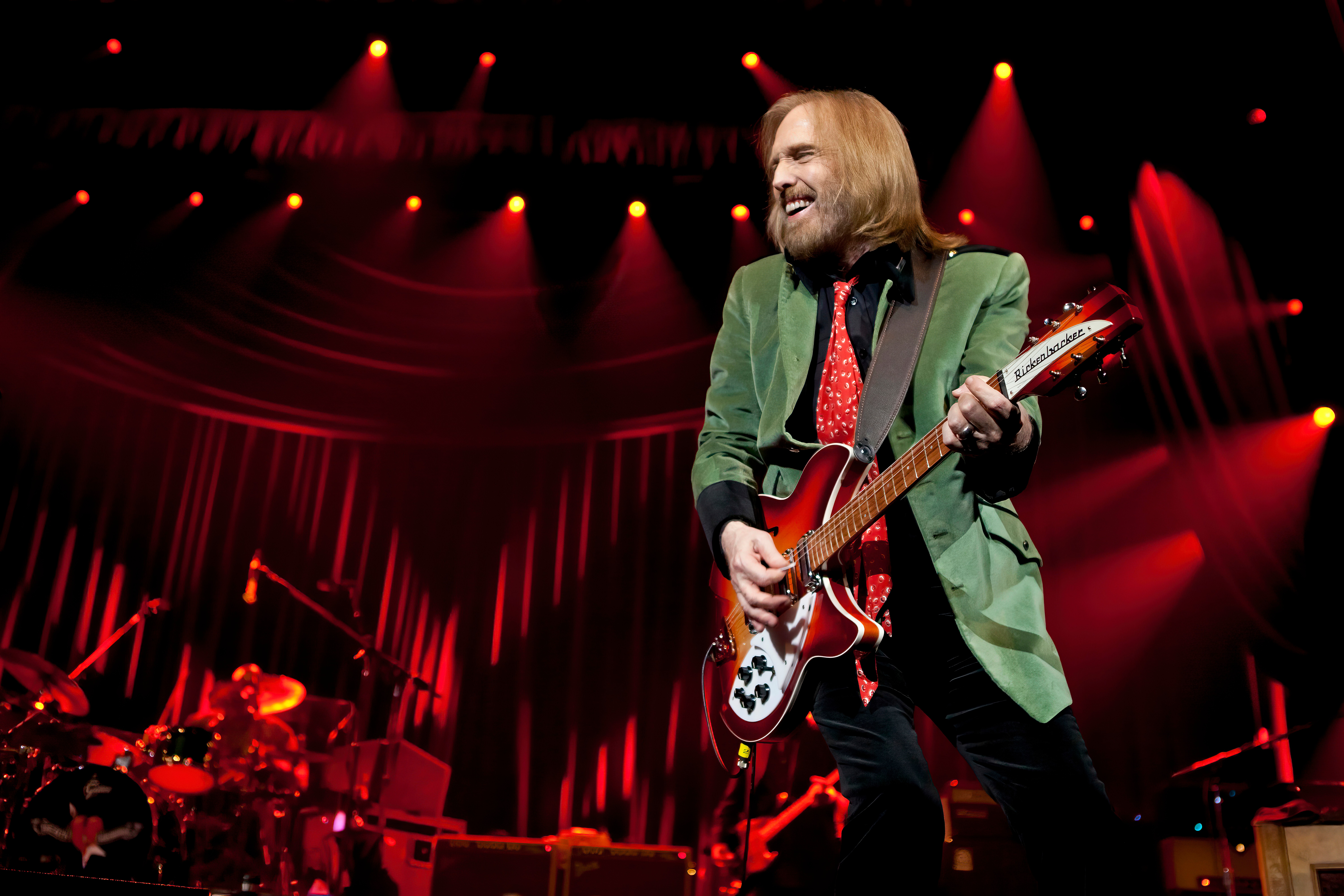 Musician with long hair and beard playing guitar on stage with red lighting and curtains behind him.
