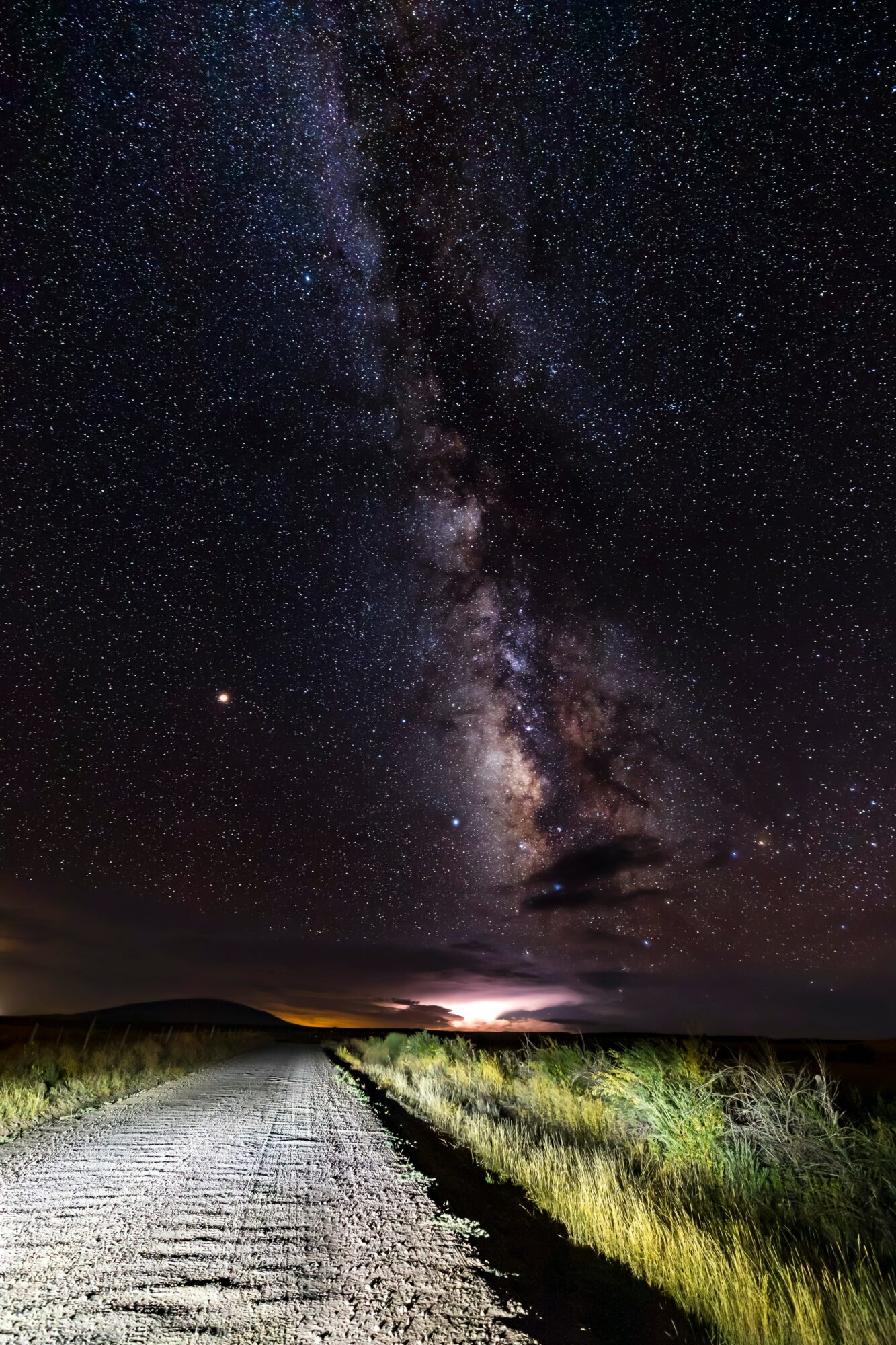 Night sky filled with stars and the Milky Way galaxy above a rural dirt road with grass on both sides.