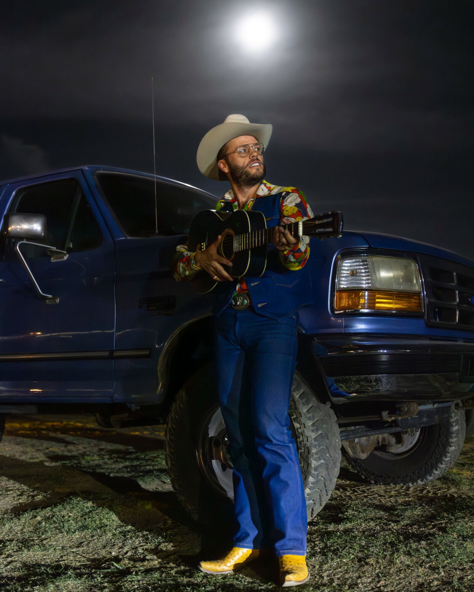 Man wearing a cowboy hat and colorful shirt playing guitar next to a blue truck under moonlit sky.