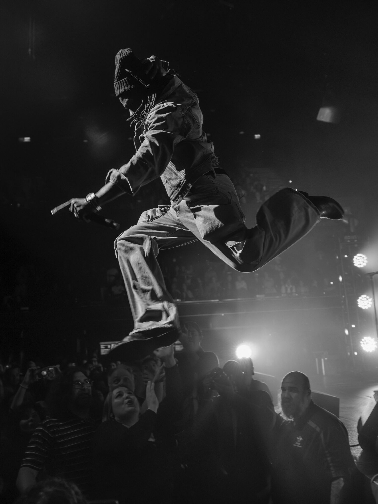 Person mid-air performing a dance move on stage with audience in foreground, black and white image.