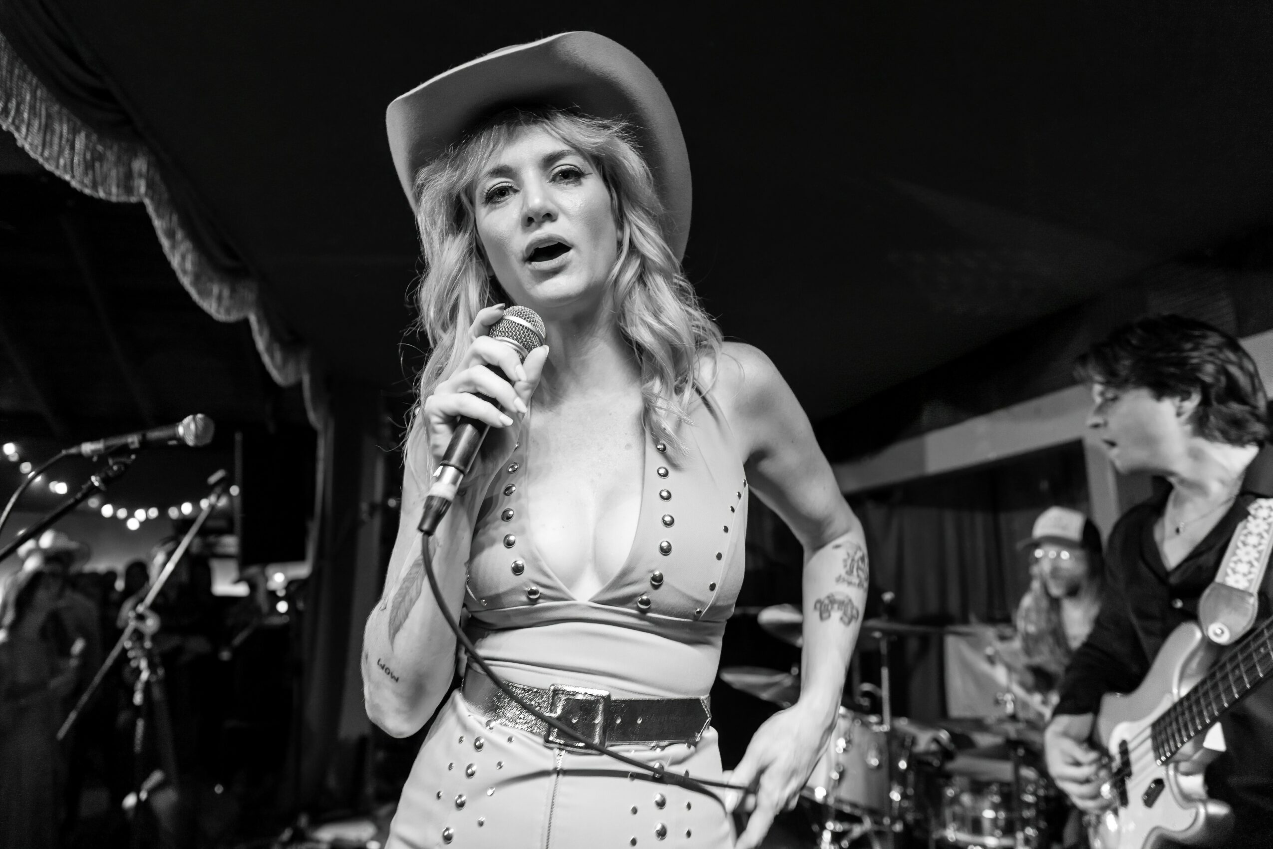 Woman singing into a microphone on stage, wearing a hat and a revealing top, with band members in background.