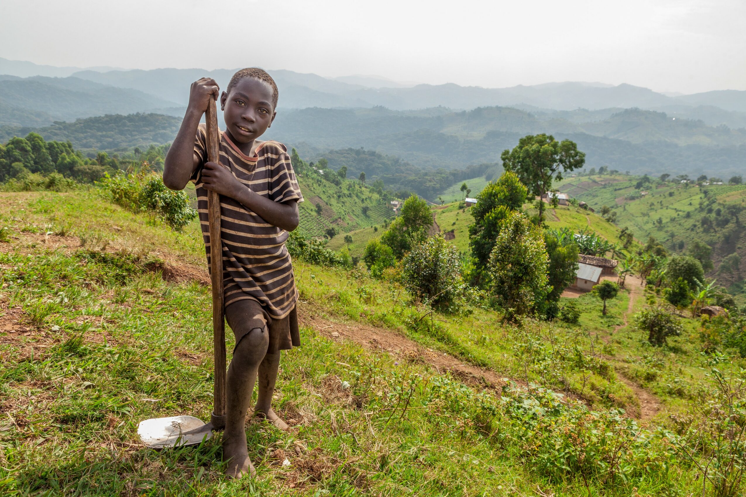 Child standing outdoors on a hillside with a walking stick, surrounded by green landscape and distant mountains.