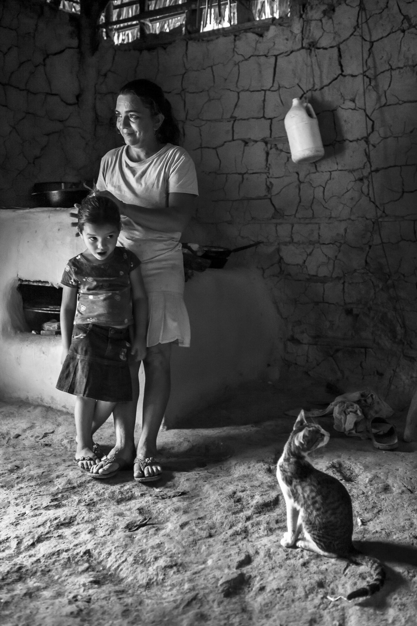 Two children standing outside near a wall with a cat sitting nearby, in black and white.