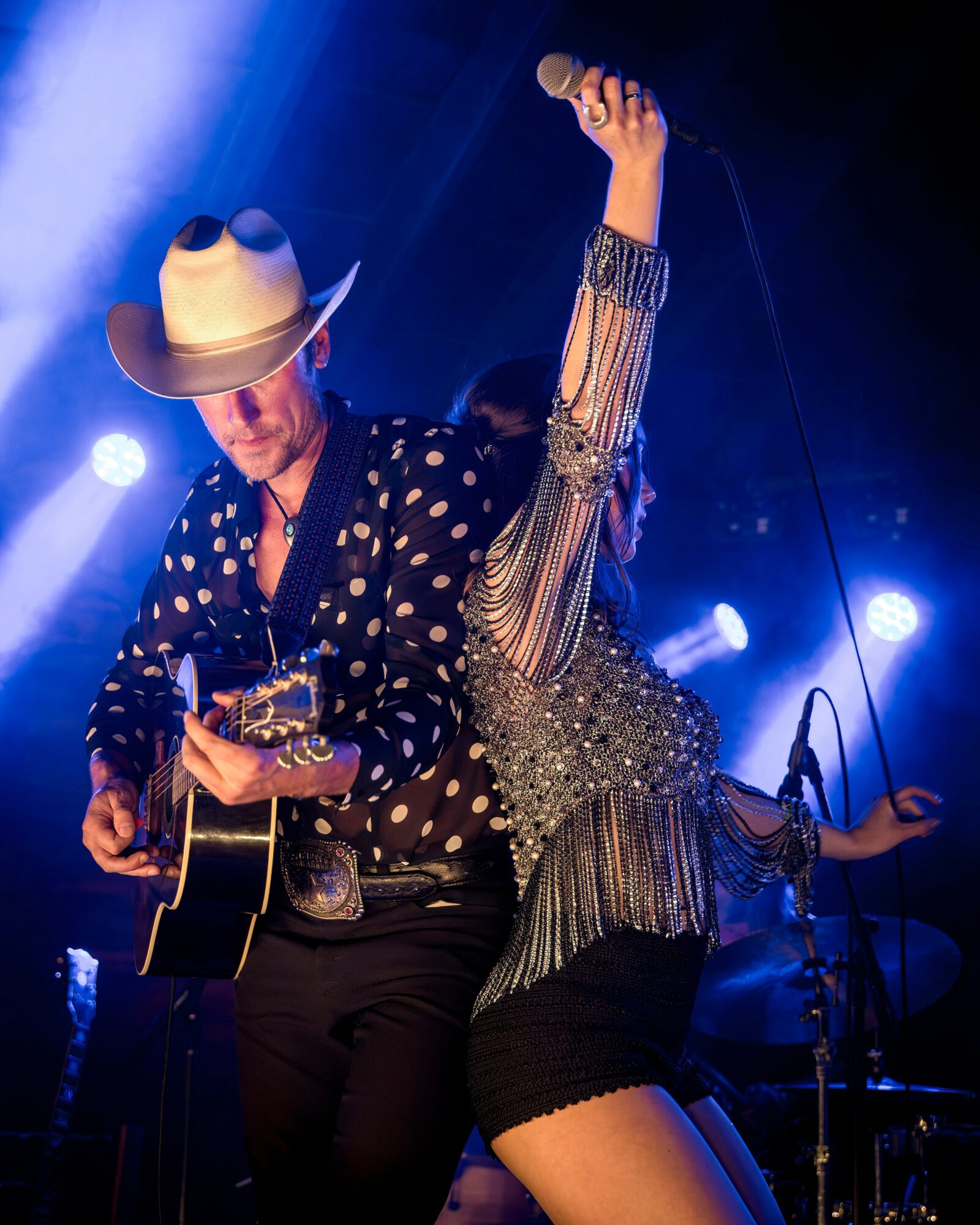 Musicians perform on stage with one playing guitar and the other raising a microphone, illuminated by stage lights.