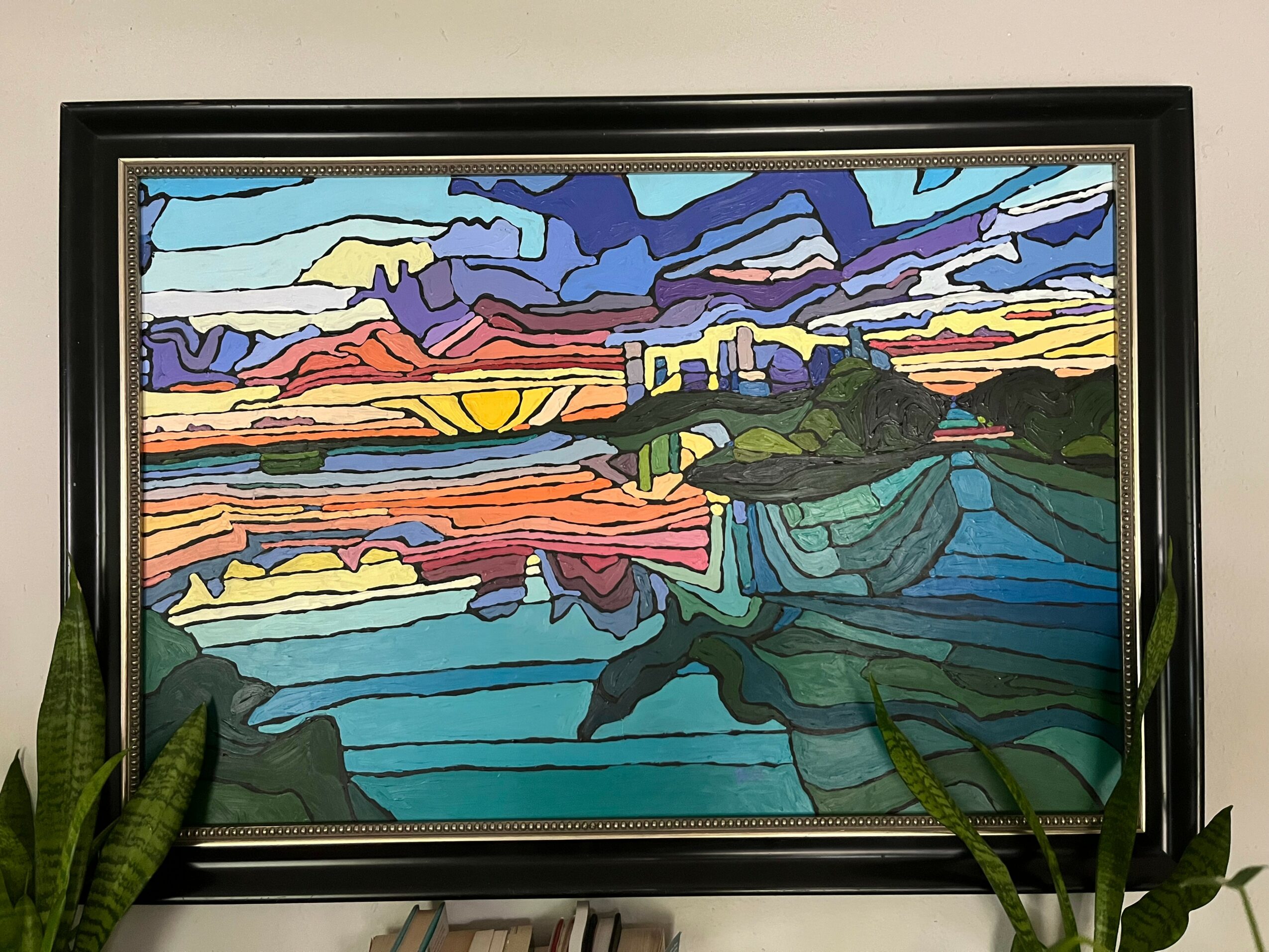 Colorful landscape painting with sky, water, and land, framed and leaning against a wall, with green plants nearby.