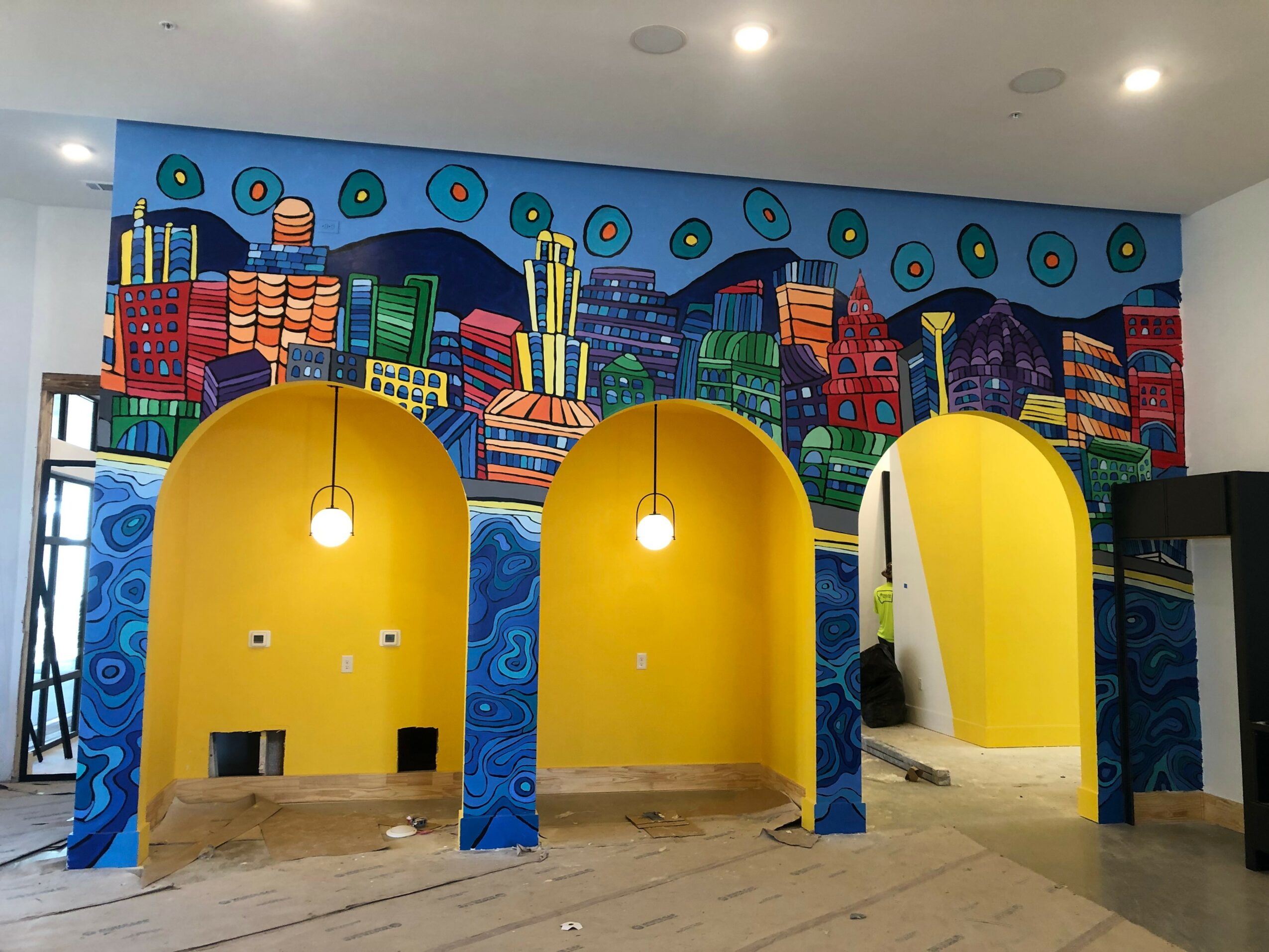 Colorful mural of a cityscape with buildings and abstract shapes, three yellow booths with hanging lights, and a wooden floor.