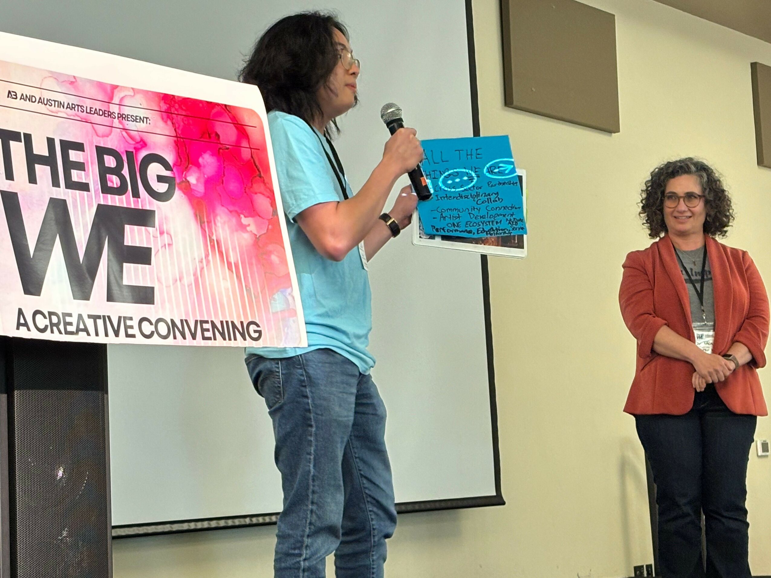 Two children standing in a room, one holding a blue book, and a sign with pink and black text on a table.