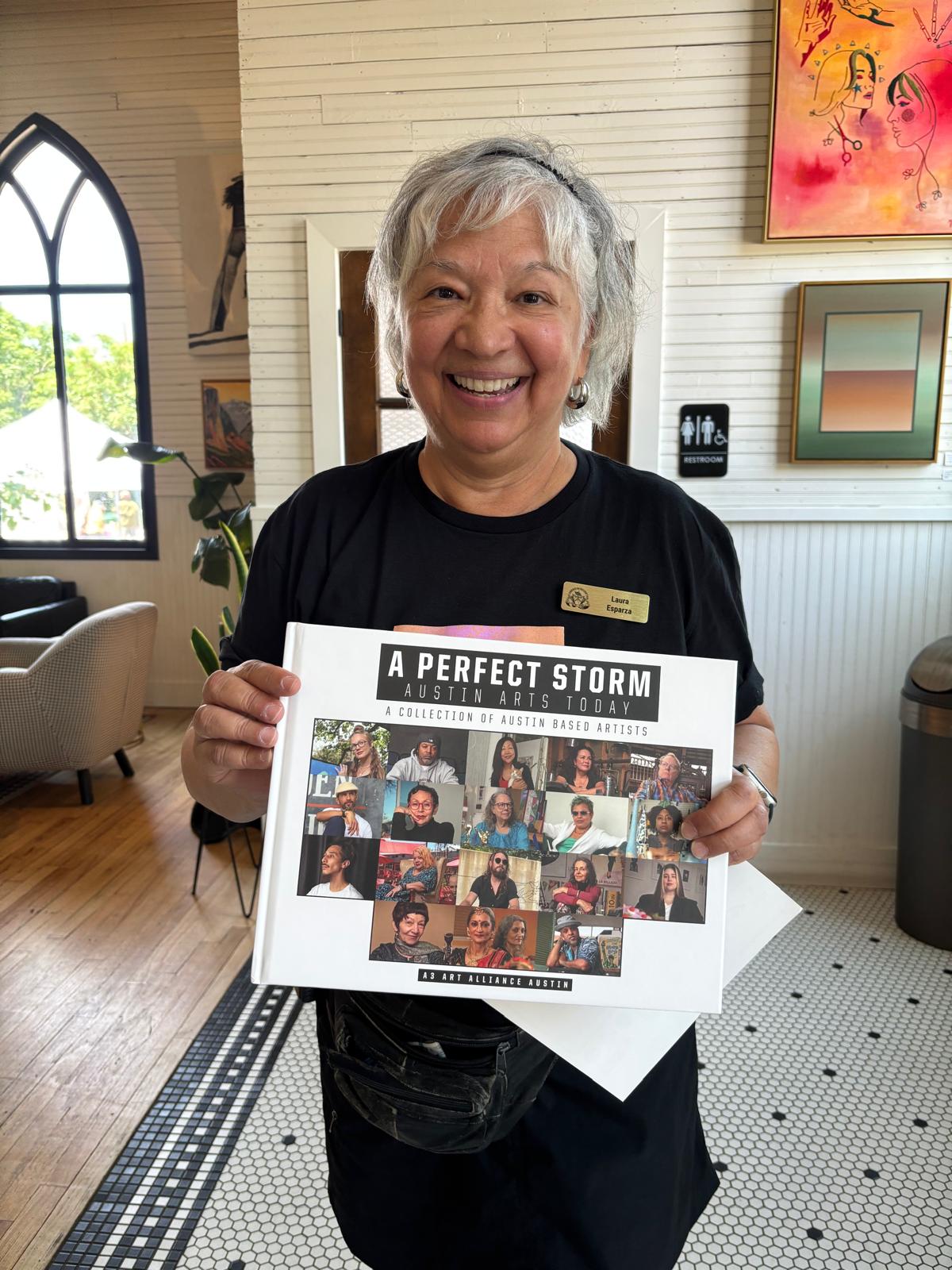 Smiling woman holding a collage of photos, standing indoors with a window and artwork in the background.
